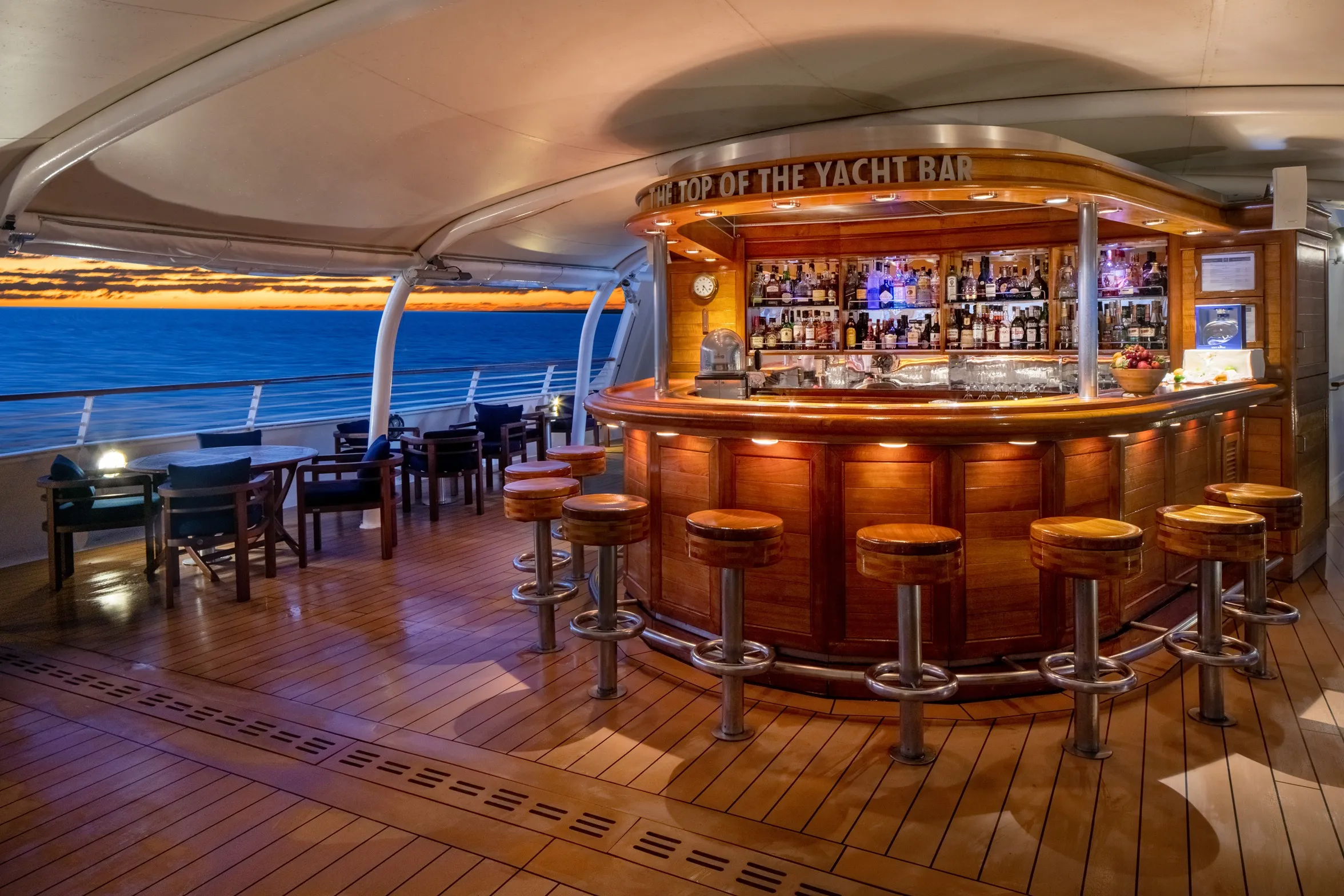 Luxurious yacht bar with wooden stools and a stocked counter overlooking the ocean at sunset.