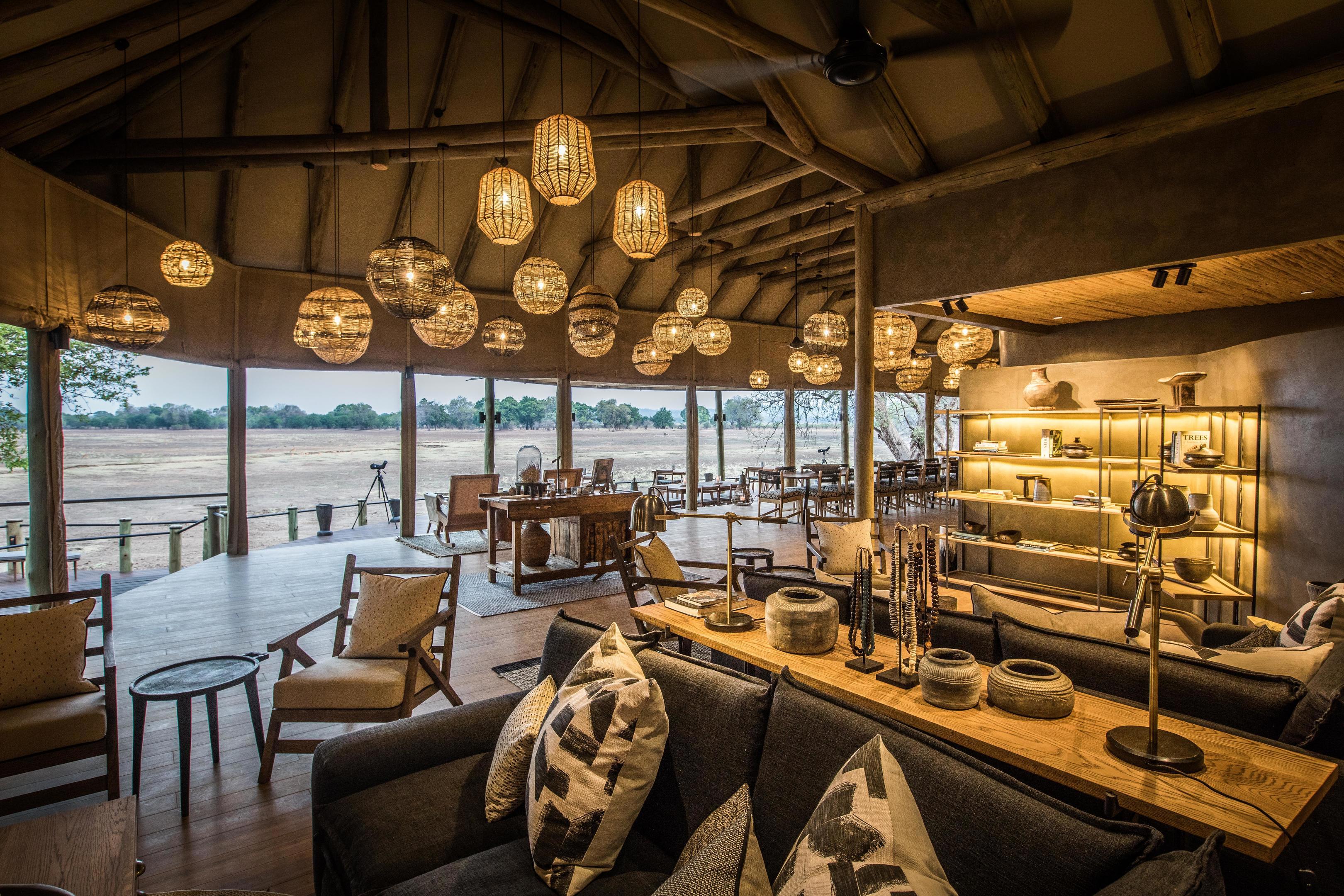 Interior of Puku Ridge Camp showcasing a spacious lounge with modern decor, wooden furniture, and large windows overlooking the landscape, illuminated by hanging lanterns for a warm ambiance