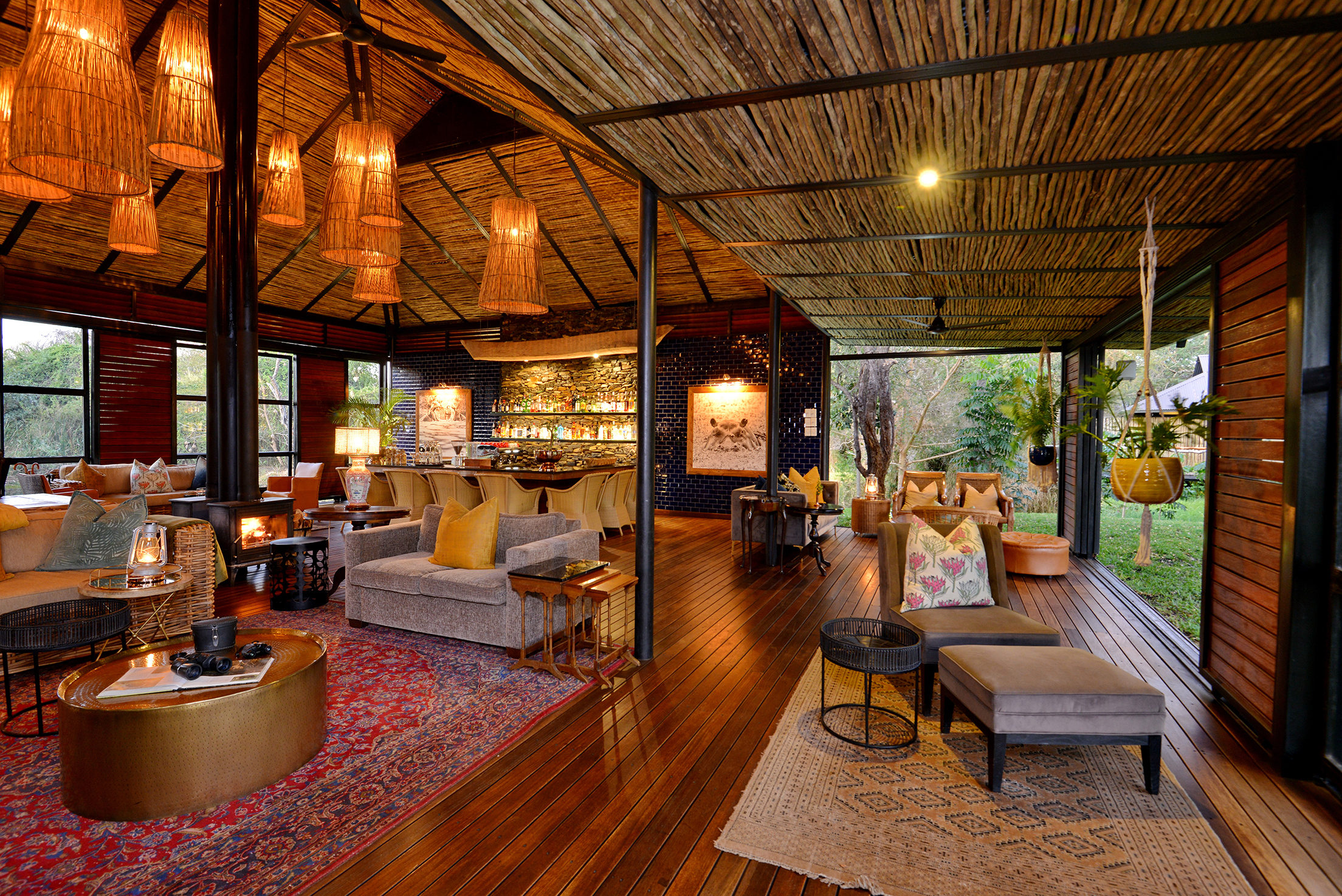 A cosy and elegant lounge area at Mukwa River Lodge with wooden floors, wicker ceiling lights, plush seating, and a well-stocked bar in the background.