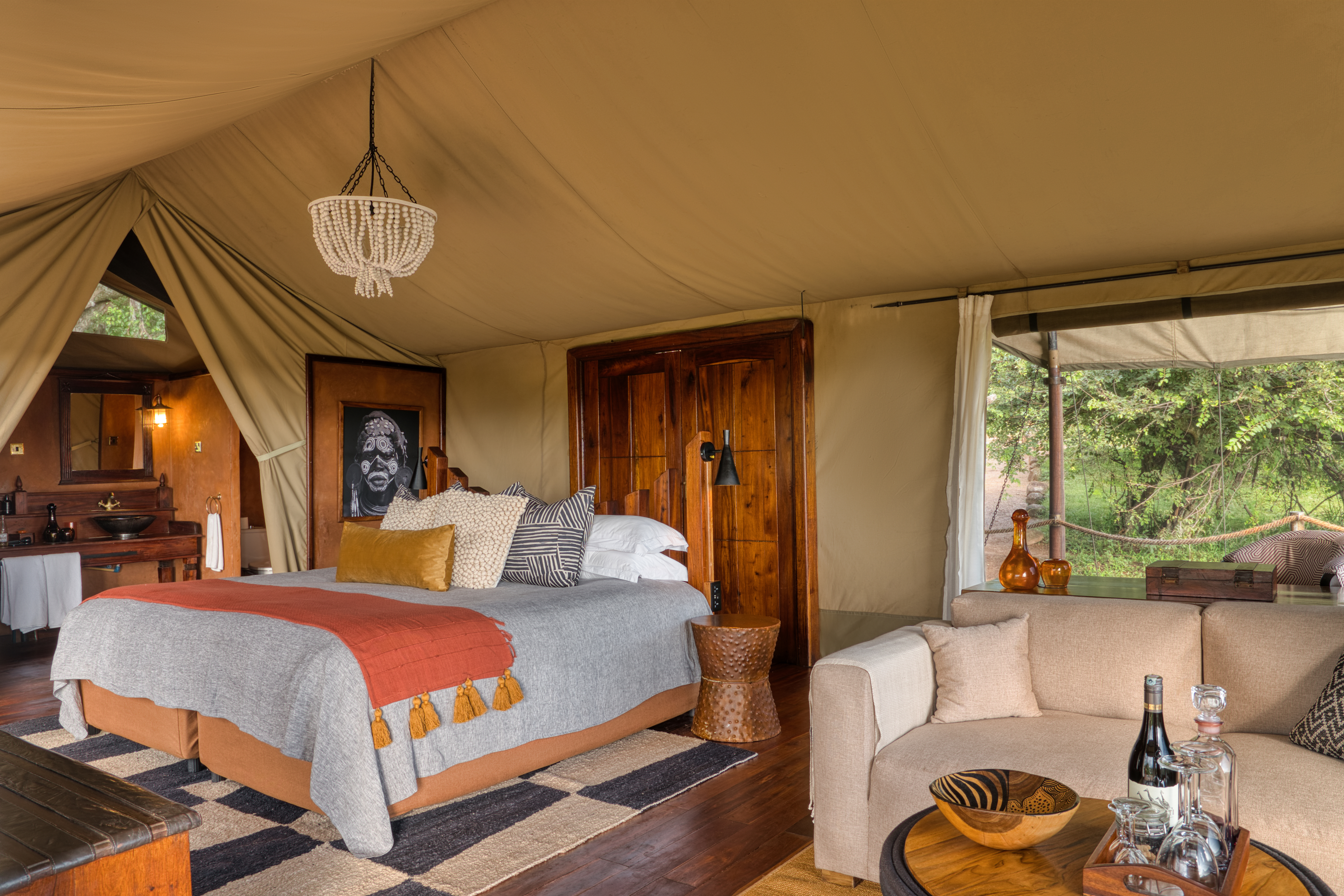 The interior of a luxury safari tent at Elewana Serengeti Migration Camp featuring plush furniture, soft furnishings in earthy tones and a beaded chandelier with glimpses of a bathroom and outside greenery