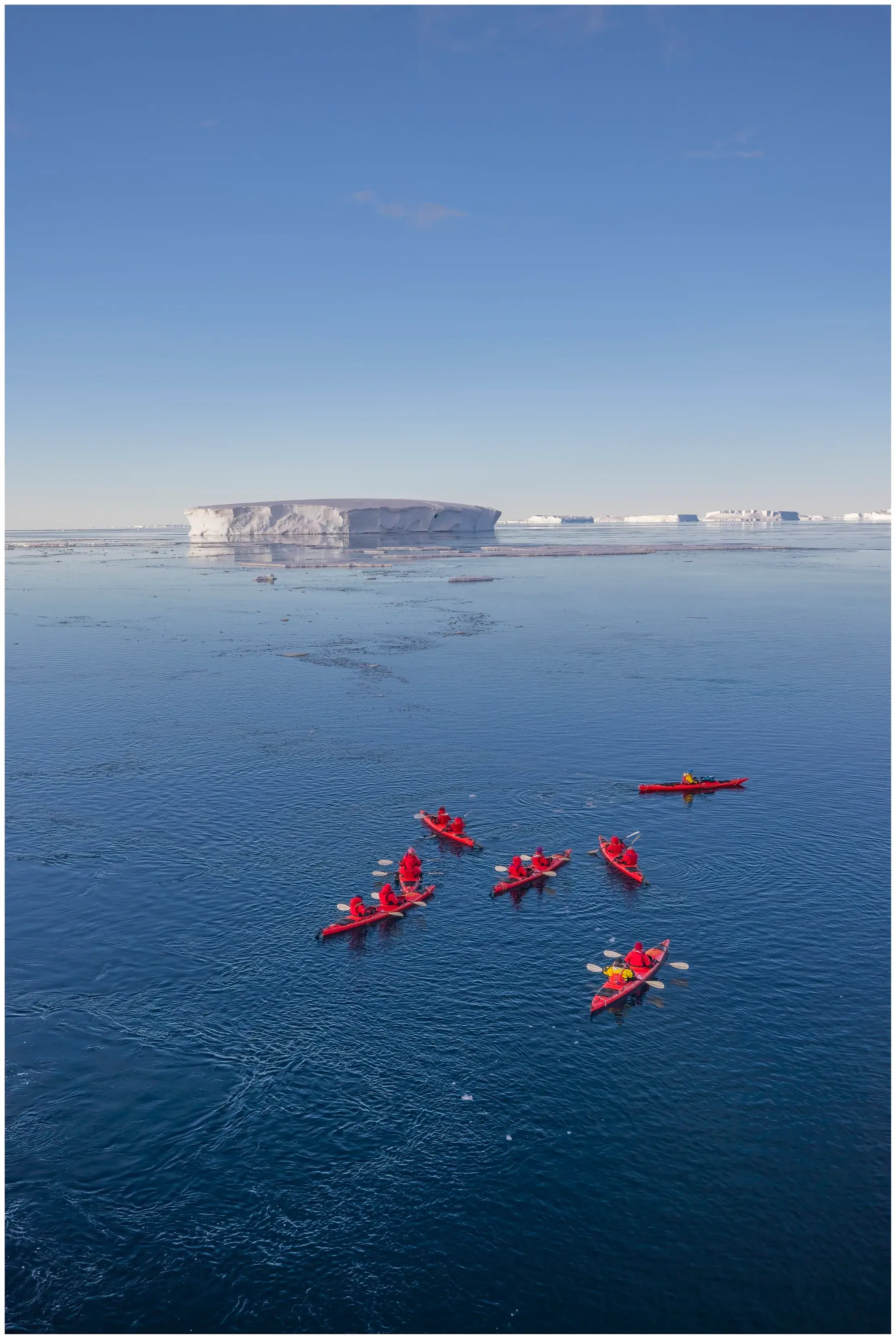 Ponant guests kayaking in icy Antarctic waters with a towering iceberg in the background under a clear sky.