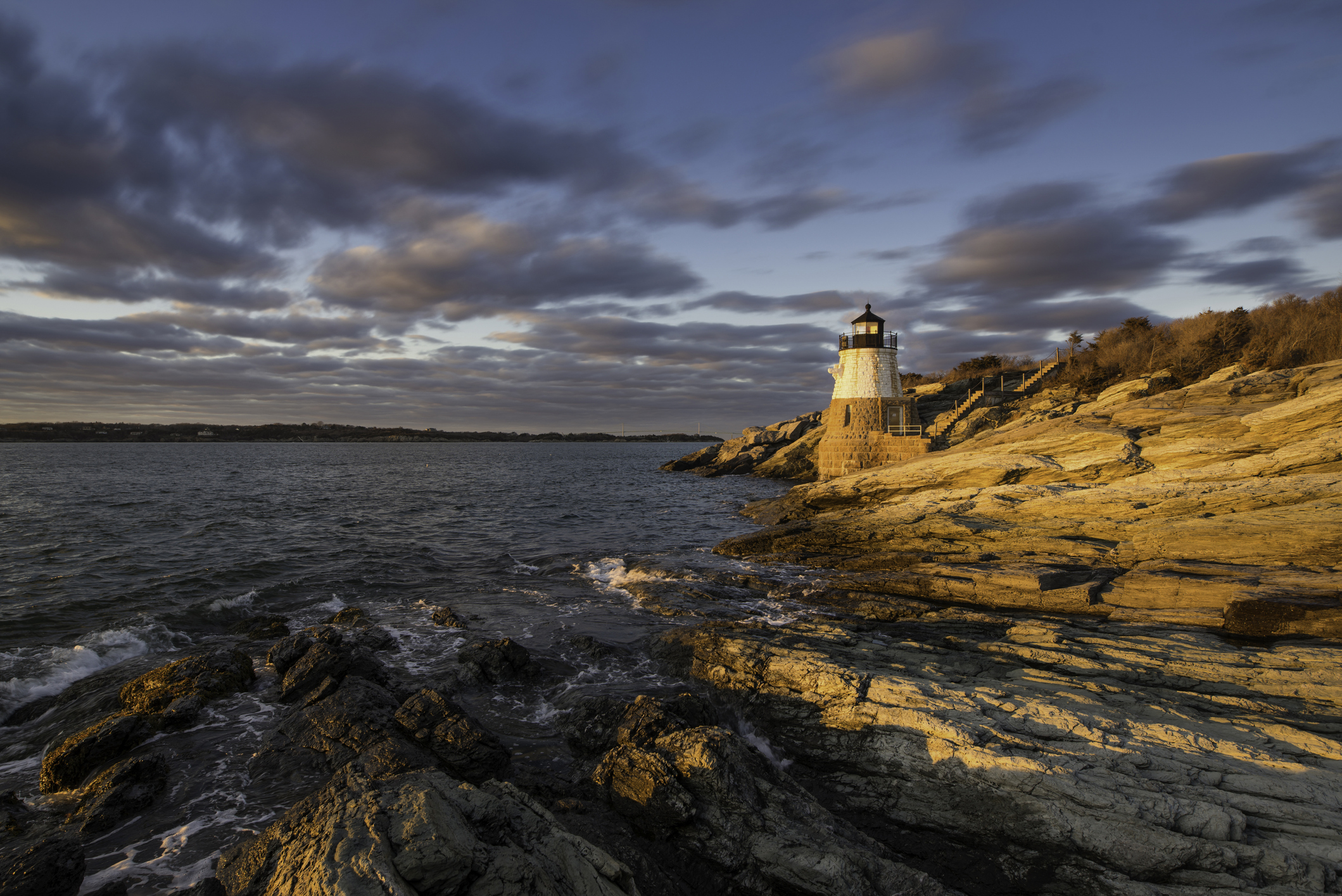 A lighthouse stands on a rocky coastline with layered sedimentary rocks, bathed in the warm glow of a setting sun under a partly cloudy sky.