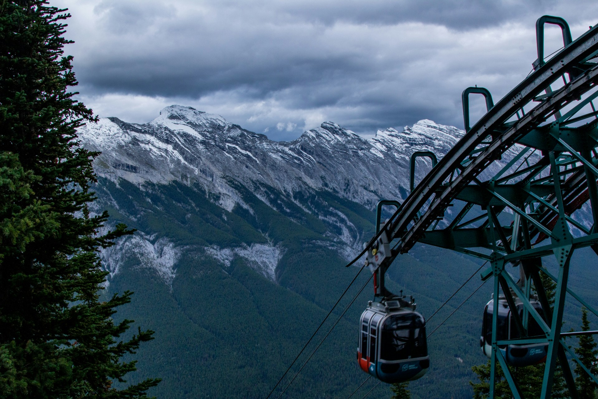 The Banff gondola lift ascending a mountain with snow-capped peaks in the background under a cloudy sky