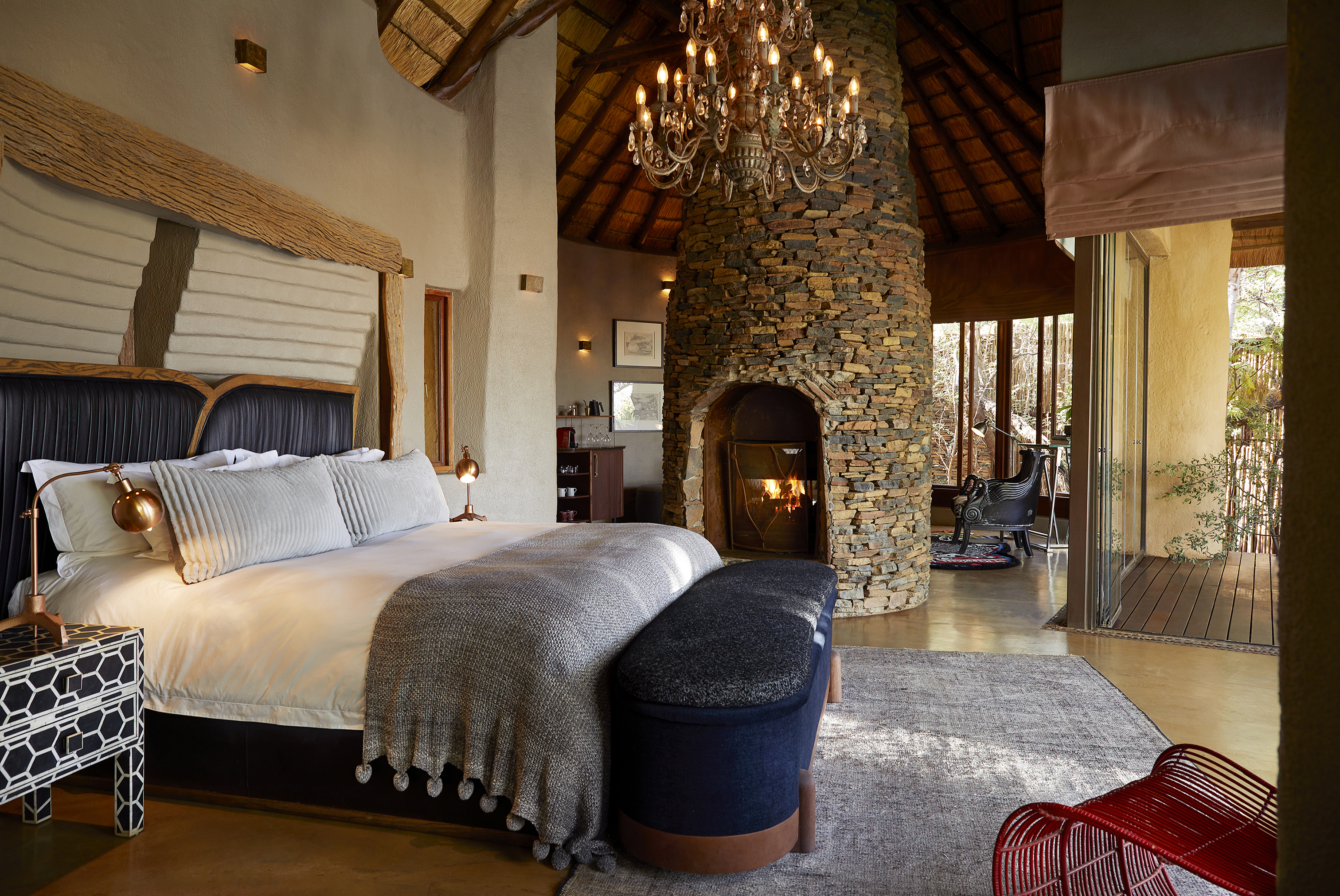 A lavish safari-style bedroom at Molori Safari Lodge with high thatched ceilings featuring a large comfy bed, furniture and a tall circular, stand-alone stone fireplace