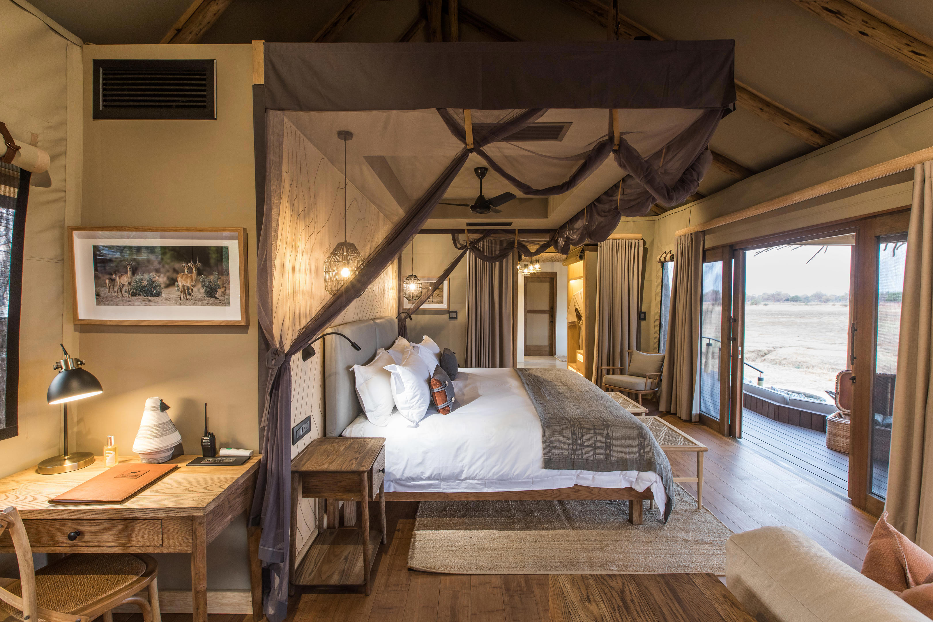 Luxurious bedroom at Puku Ridge Camp with a canopy bed, wooden furnishings, and large glass doors opening to a deck overlooking the wilderness, warmly lit and styled in earthy tones