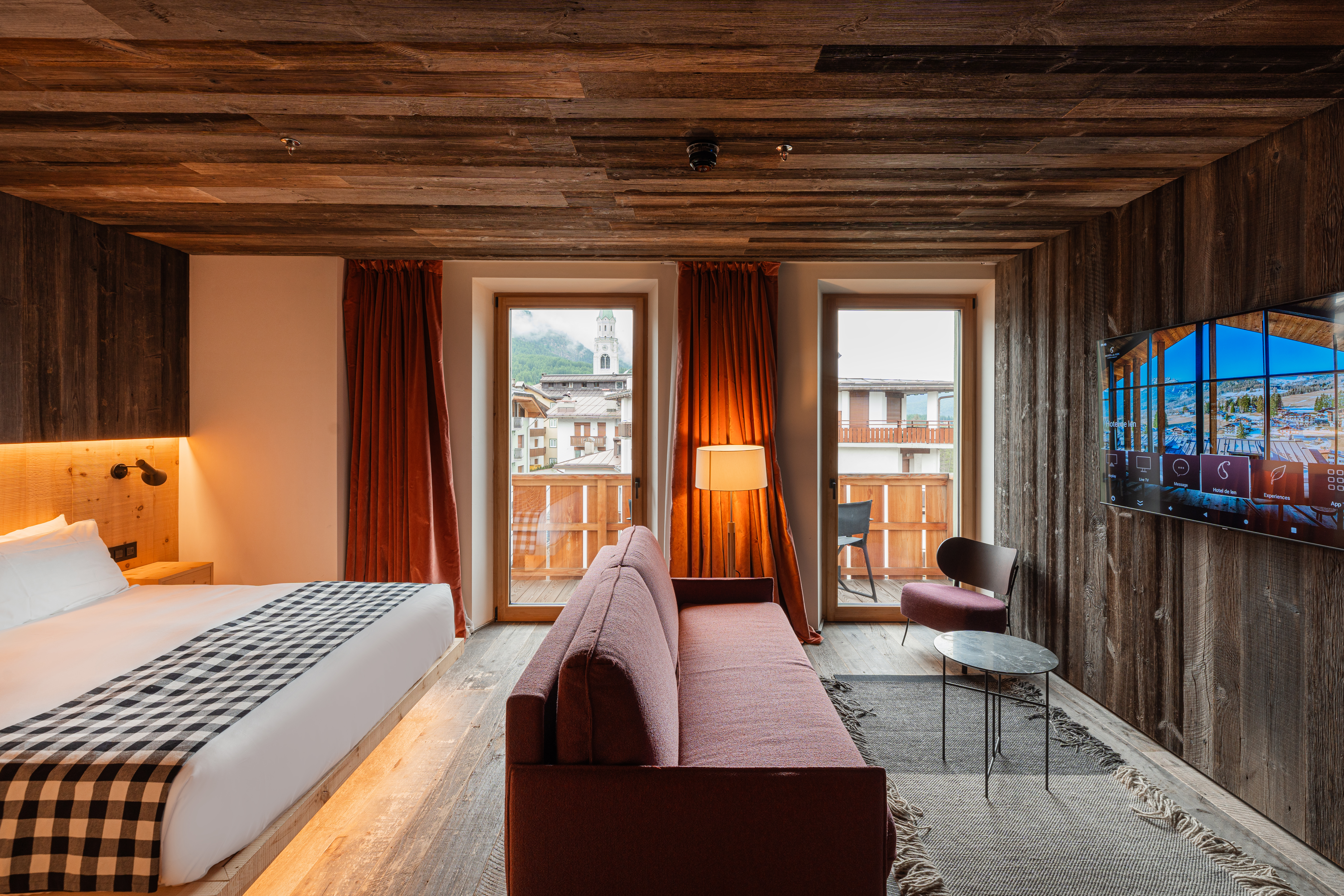 Wide shot of family room featuring a long red sofa, bed with checkered throw, television and two doors to a balcony