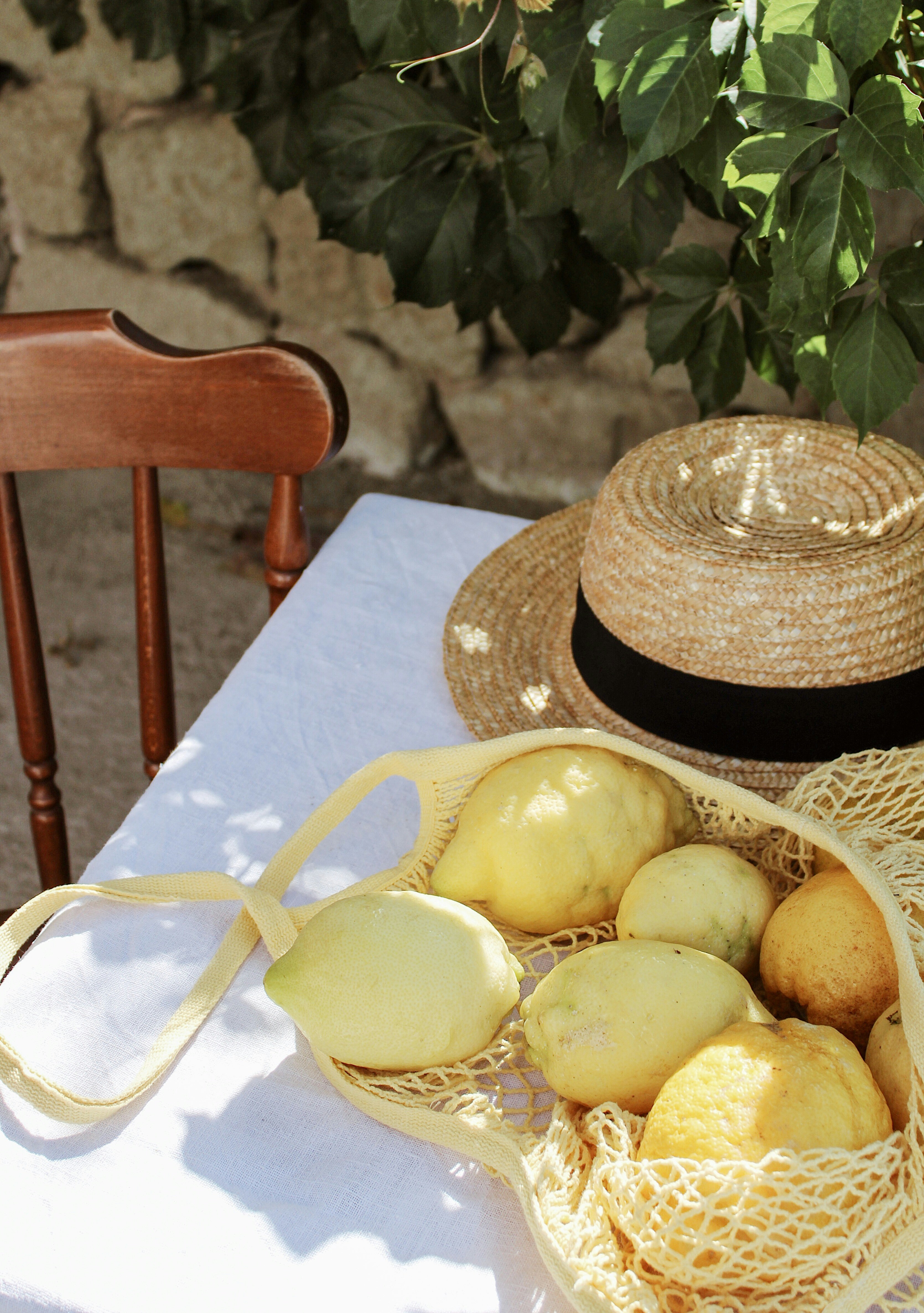 Fresh lemons in a mesh bag on a white table with a straw hat under leafy shade in a rustic outdoor setting.