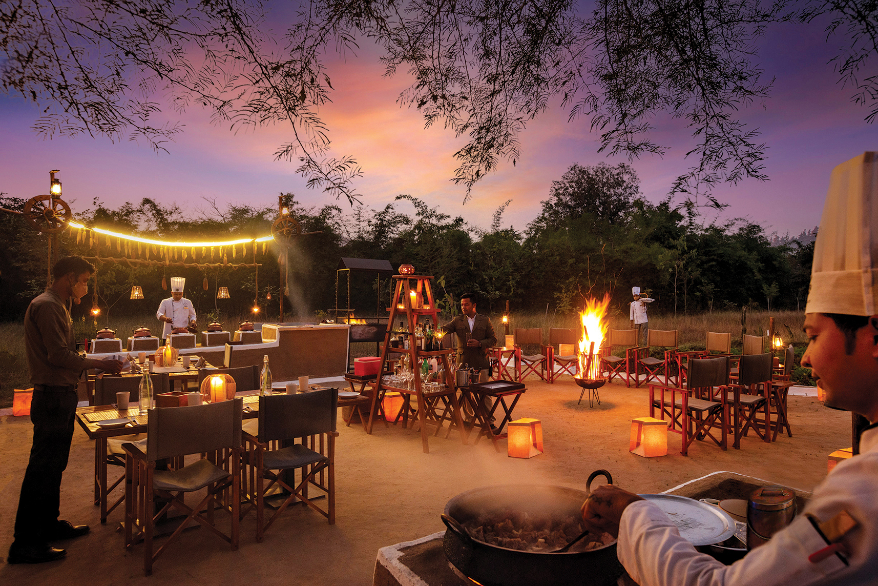 Chefs and waiters attending to duties as they set up a meal surrounding a fire pit at dusk