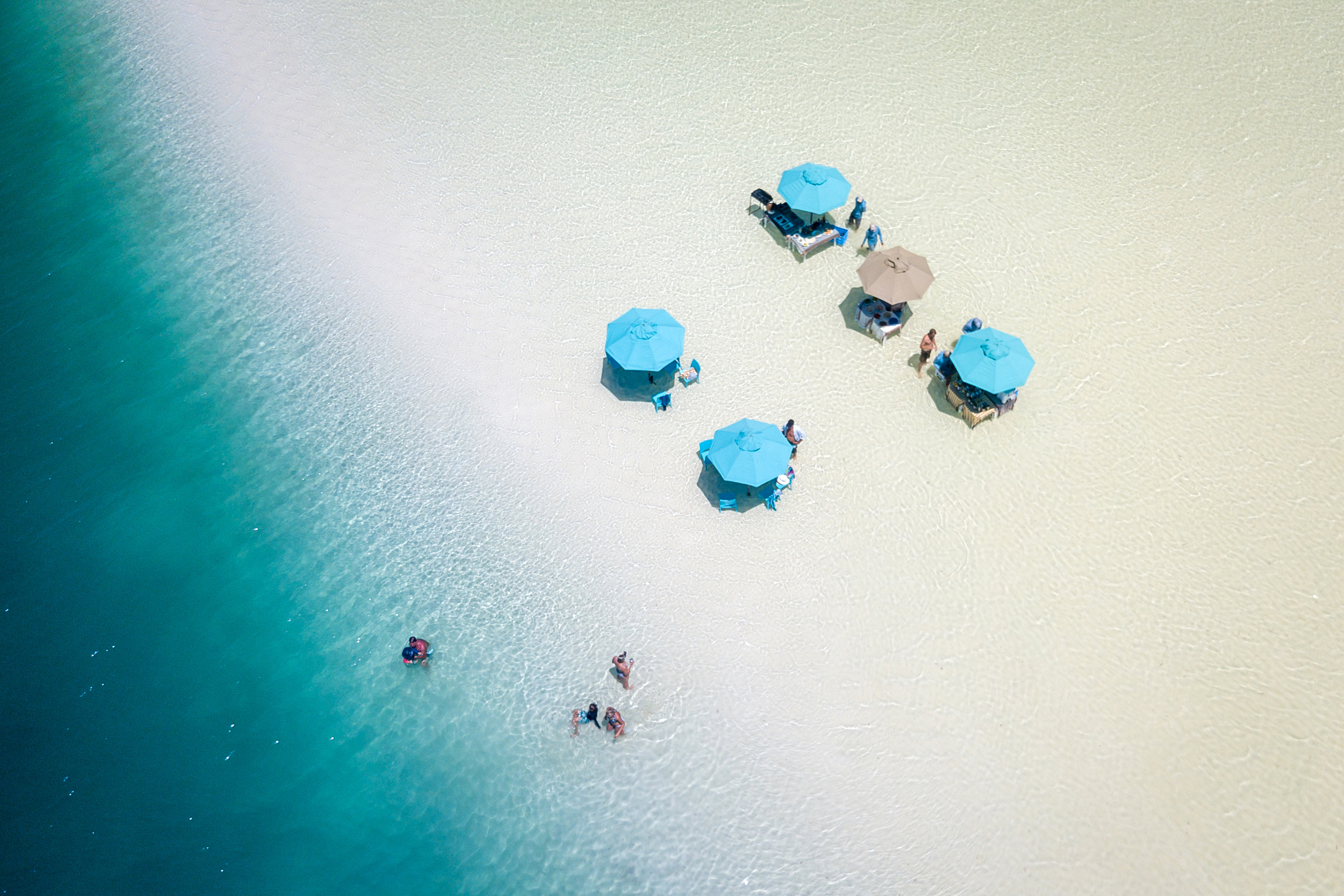 People enjoying time on the beach with tables and blue sun umbrellas set up for lunch beside the water