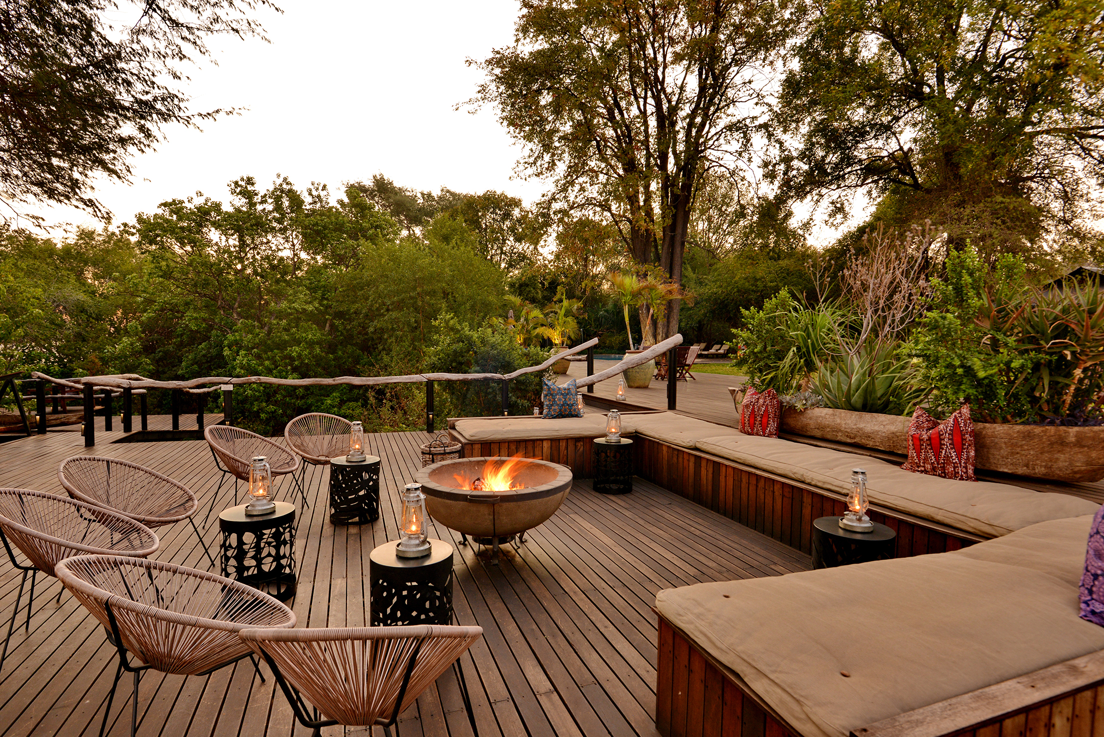 Outdoor seating area at Mukwa River Lodge with a central fire pit, wicker chairs, cushioned benches, and a wooden deck surrounded by lush greenery.