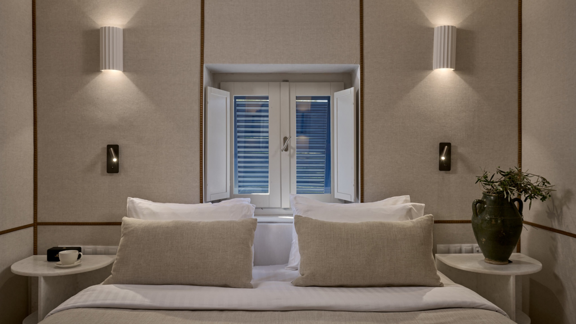 The top of a plush neutral toned bed below a small square window with shutters, flanked by tables with a vase and coffee cup and wall lights
