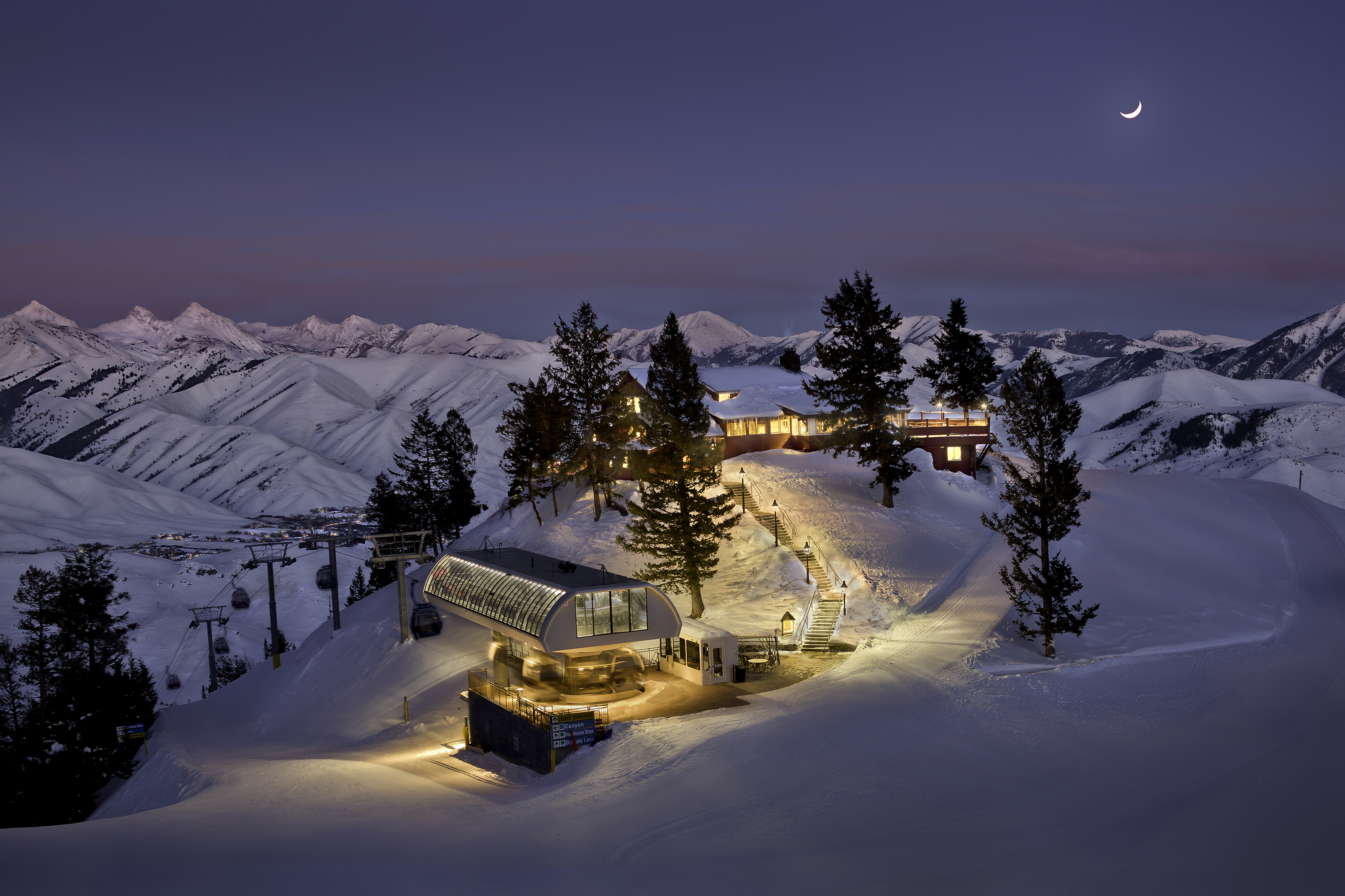 A snowy mountain landscape at dusk with The Roundhouse Restaurant and lift station illuminated by warm lights, surrounded by pine trees and snow-covered peaks