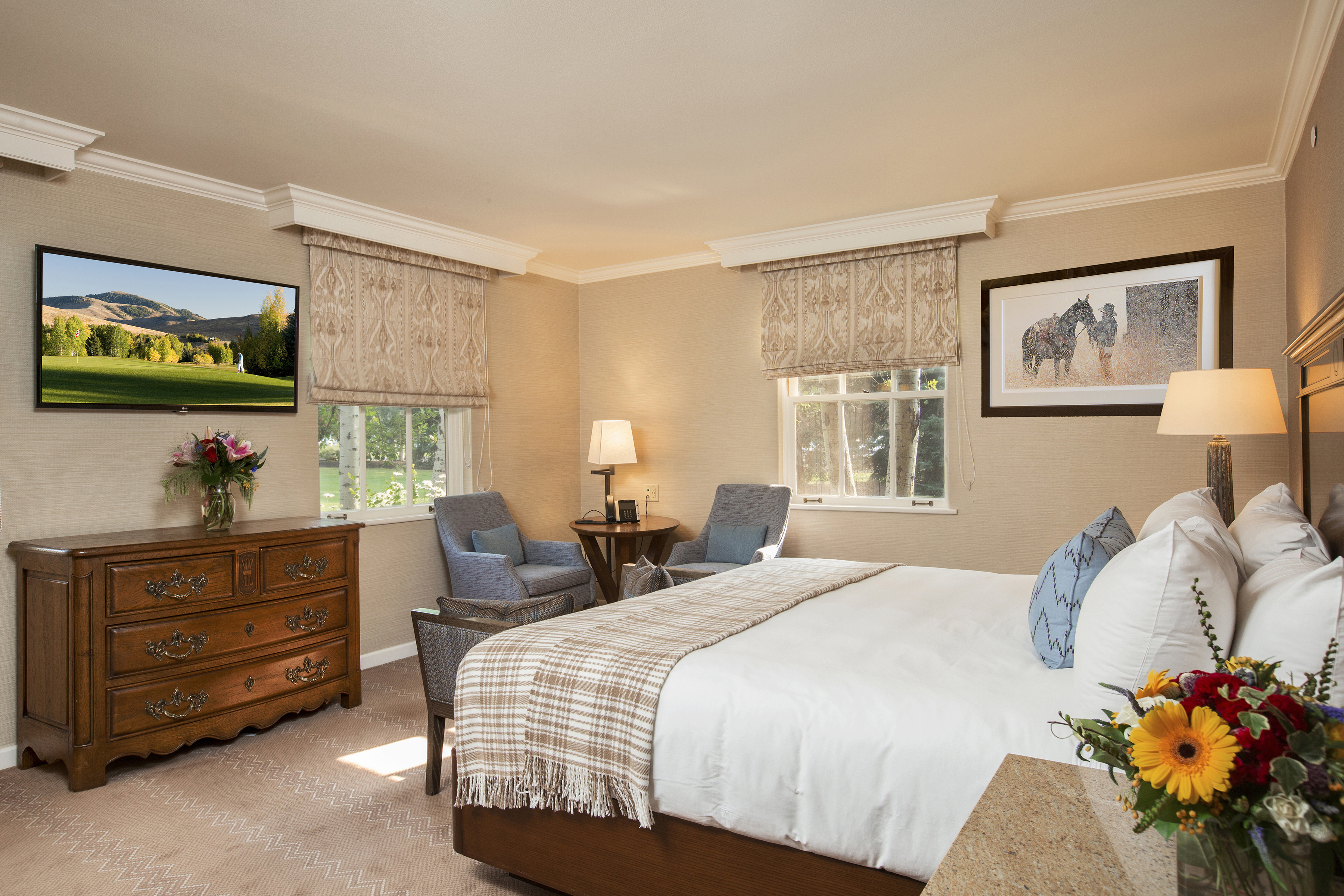 A cosy hotel room at Sun Valley Lodge featuring a large bed with white linens, two blue armchairs by the window, a wooden dresser with a vase of flowers, and framed artwork on the walls