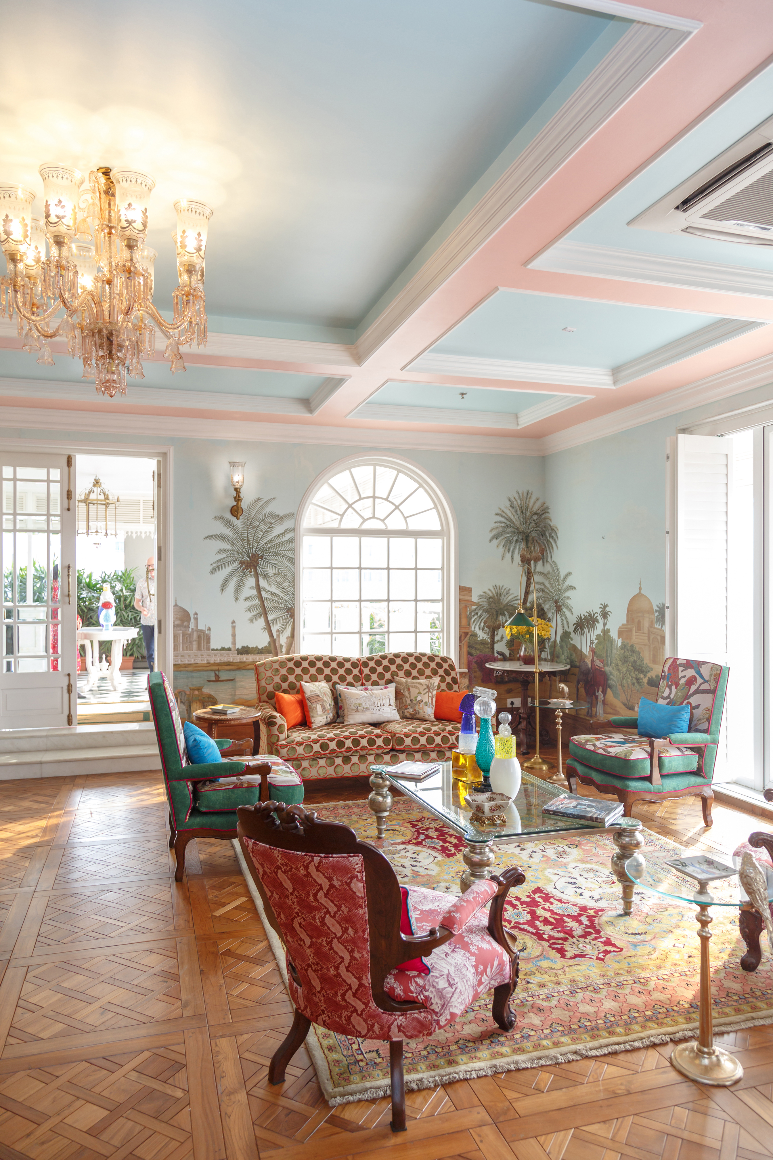 An opulent and colourful drawing room with mural painted walls, ornate furniture, chandelier and arched windows