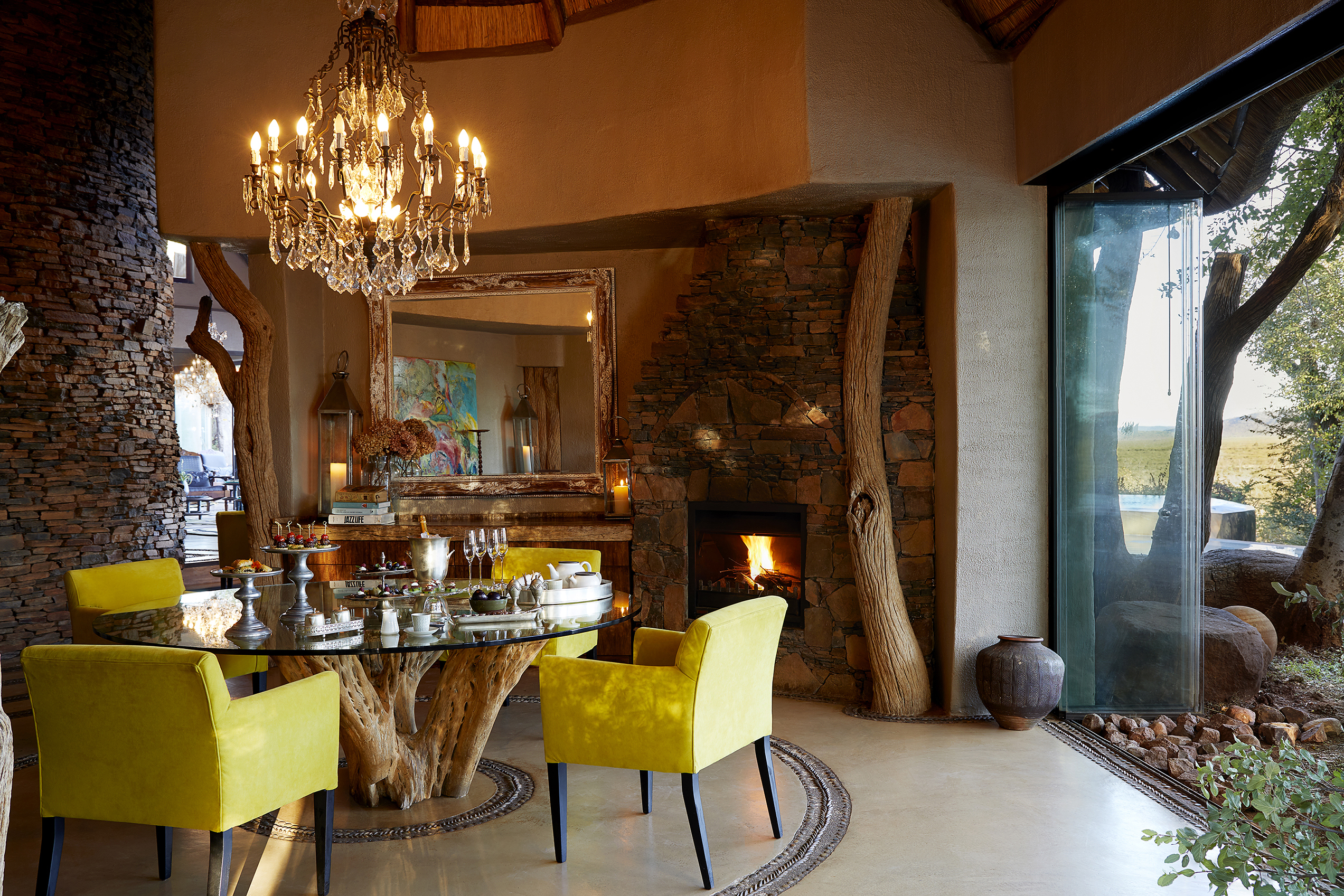 A table and yellow chairs set beneath a chandelier in the dining room of Molori Lodge beside a large window and stone wall featuring a mirror