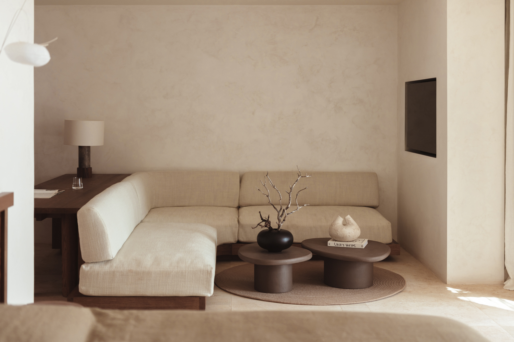 A stylish and minimal lounge area with white walls and flooring featuring a cream sofa backed by a table and wooden coffee tables topped with vases