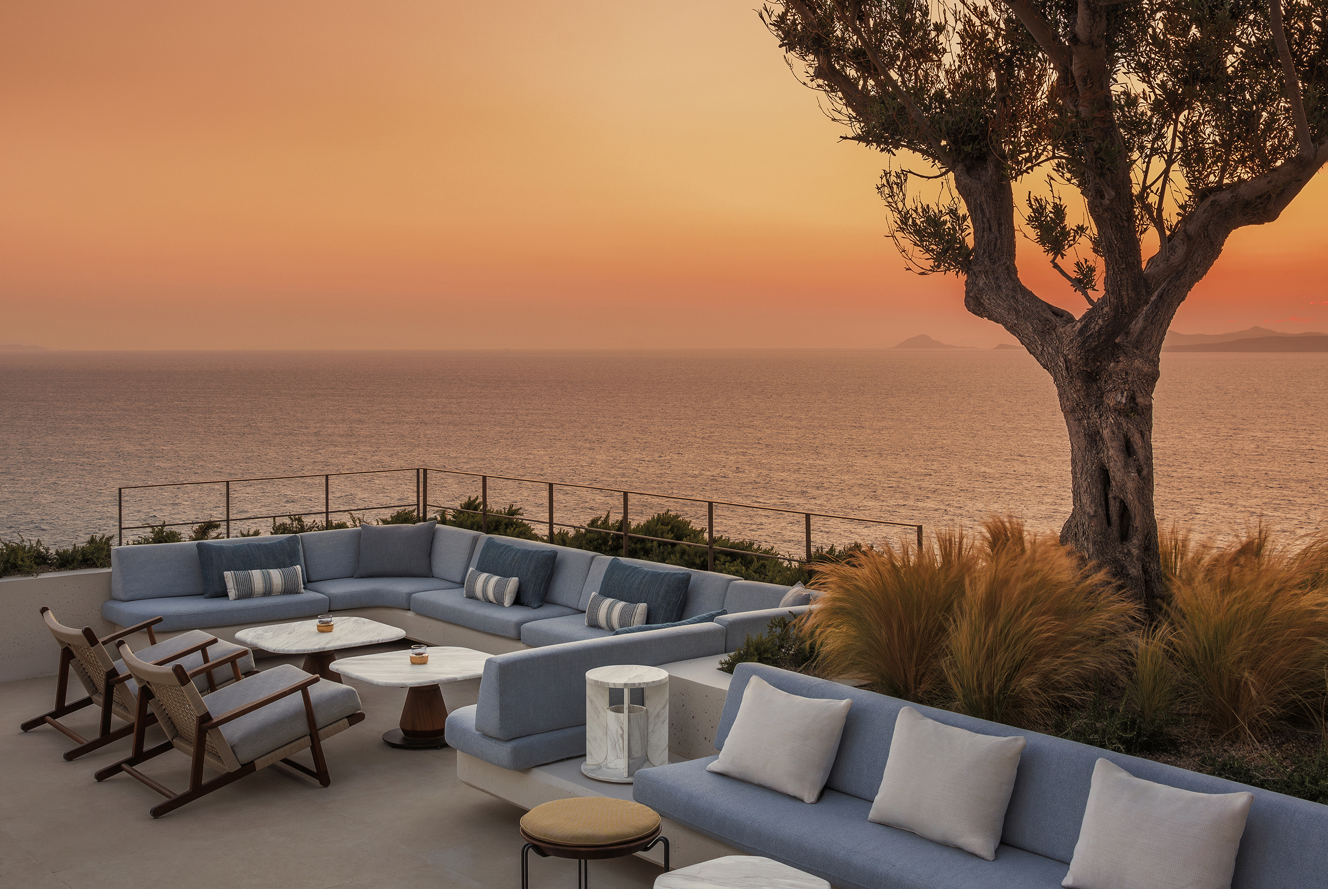 Outdoor balcony with blue cushioned sofas, marble tables and olive tree at sunset