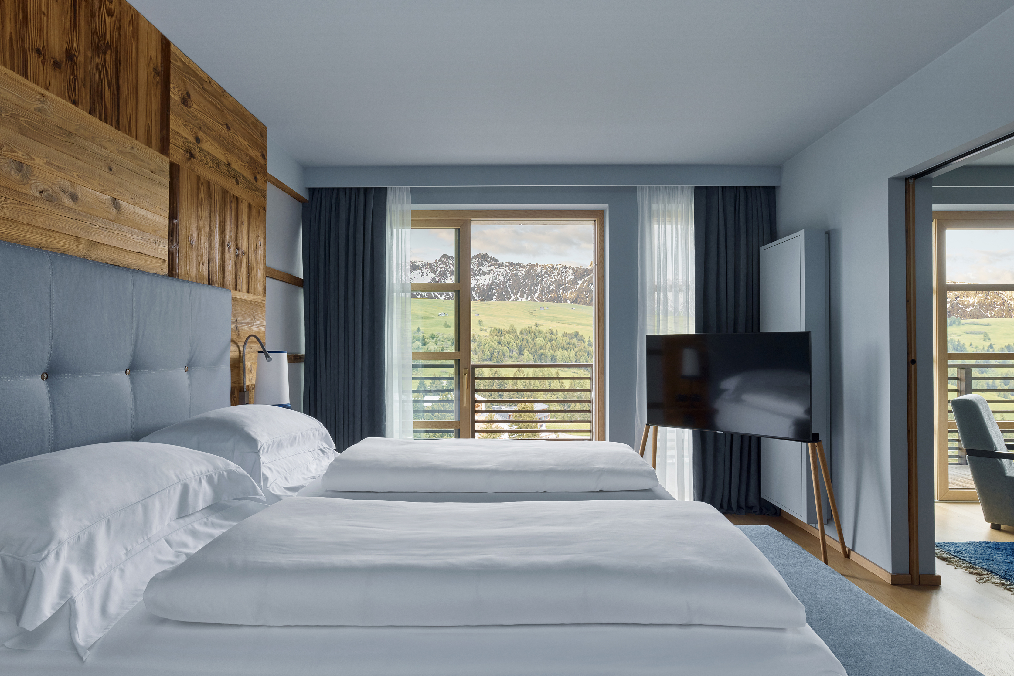 A modern bedroom with two large beds, wooden accents, and a scenic view of rolling hills through the balcony doors.