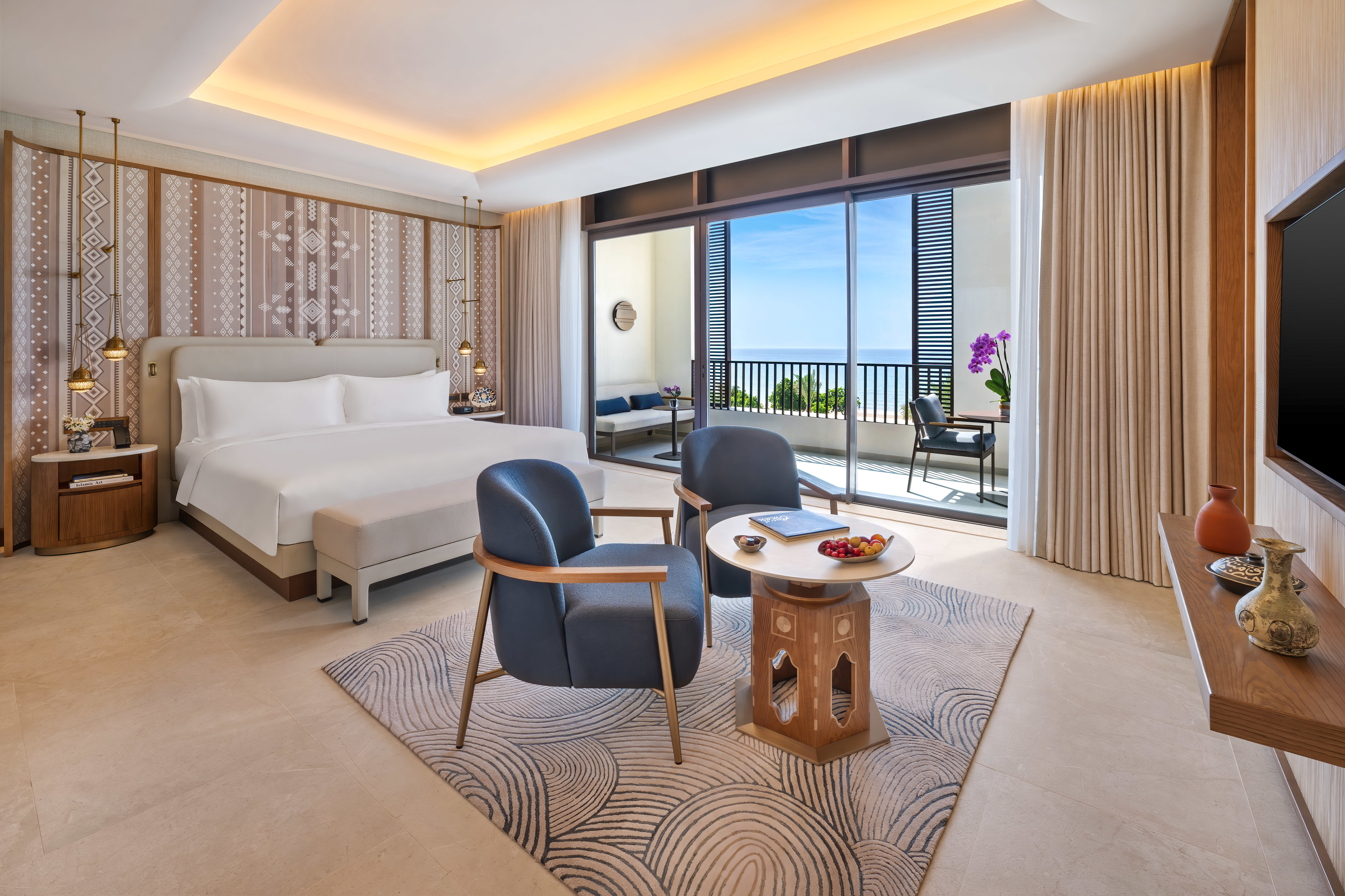 A sleek hotel suite at Mandarin Oriental Muscat featuring plush blue chairs, a comfy bed and local soft furnishings