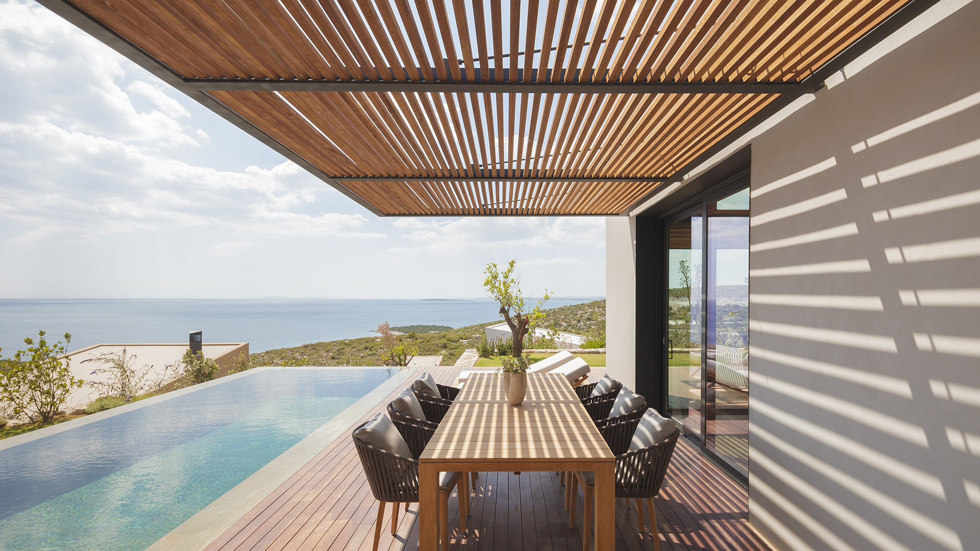 A decked area outside a private residence with a long table and chairs is flanked by an outdoor pool