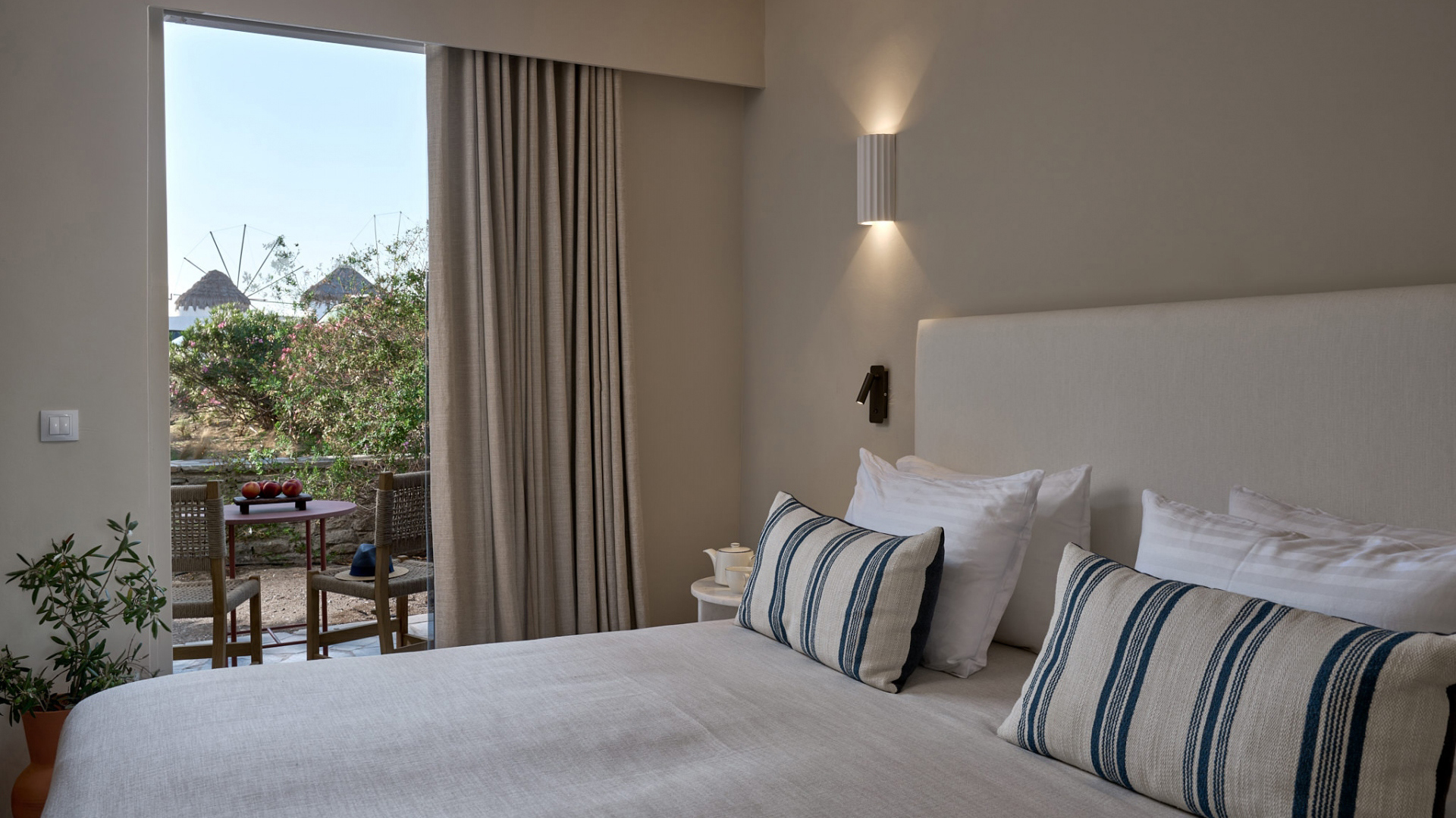 A comfy bed in neutral tones featuring blue striped cushions beside an open door with a view of greenery and the iconic Mykonos windmills