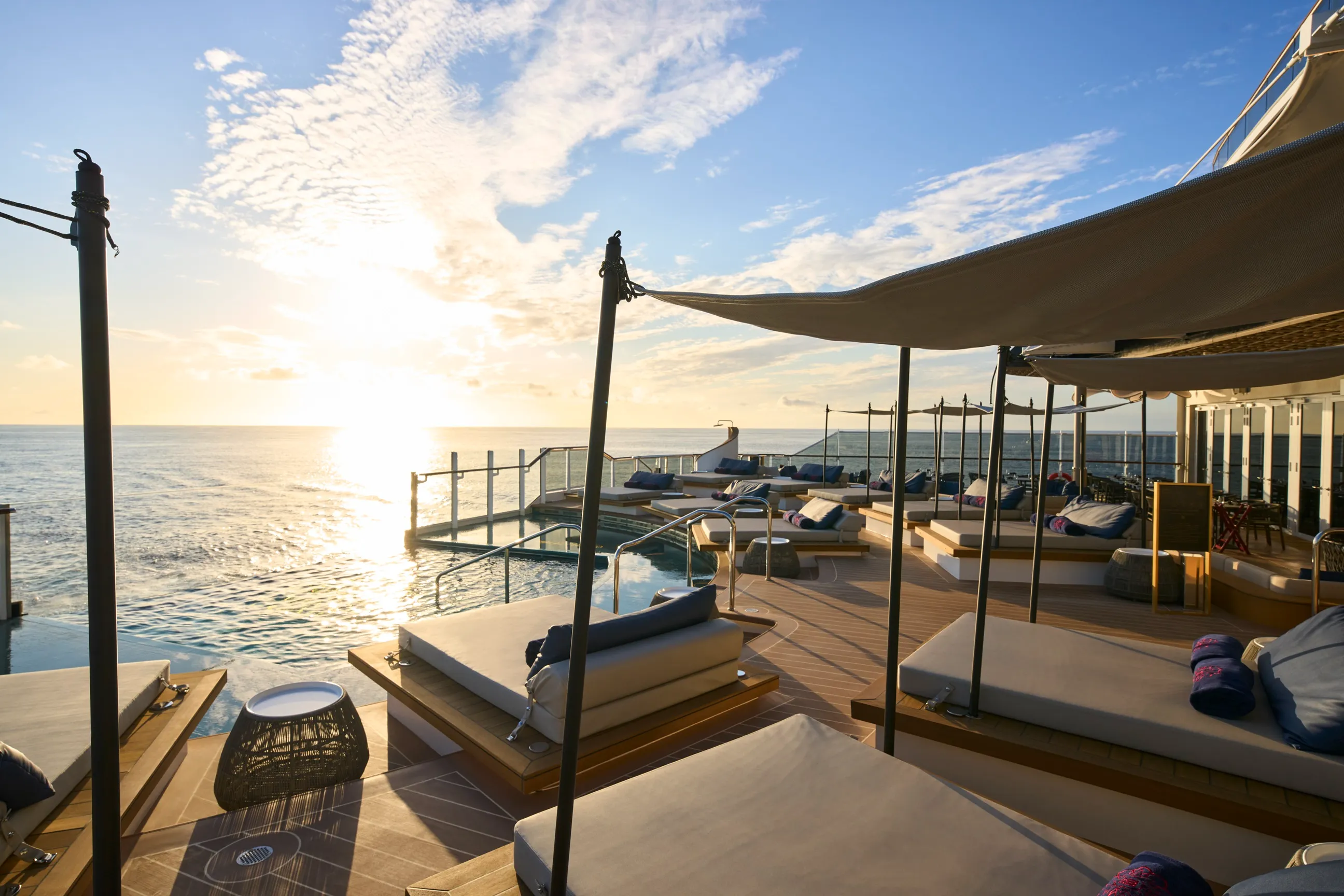 Stylish cruise ship lounge with cushioned sunbeds and canopies, set against a serene ocean sunset.