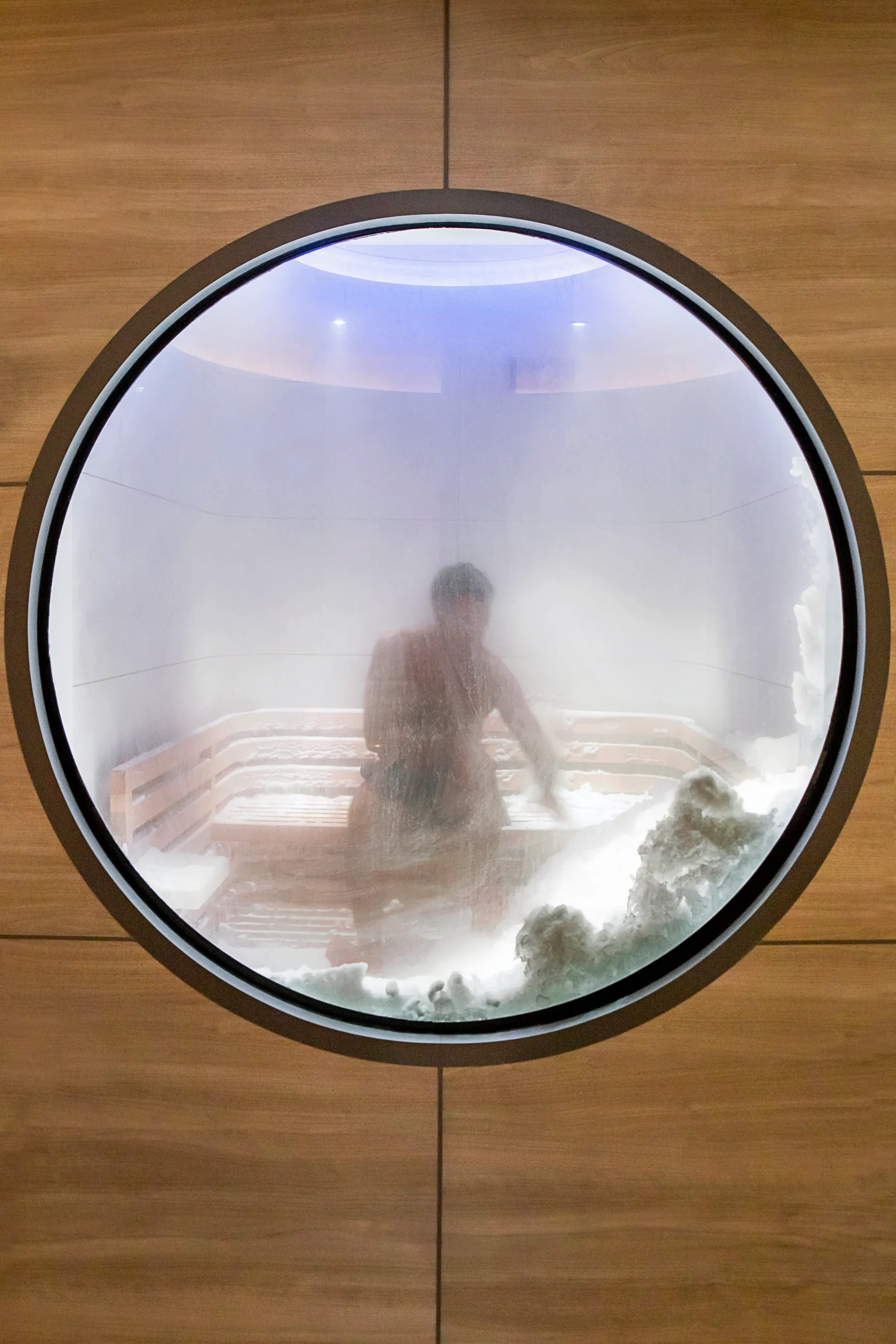 Guest seated in a snow room aboard Ponant’s Le Commandant Charcot, seen through a round foggy window, surrounded by mist and wooden benches.