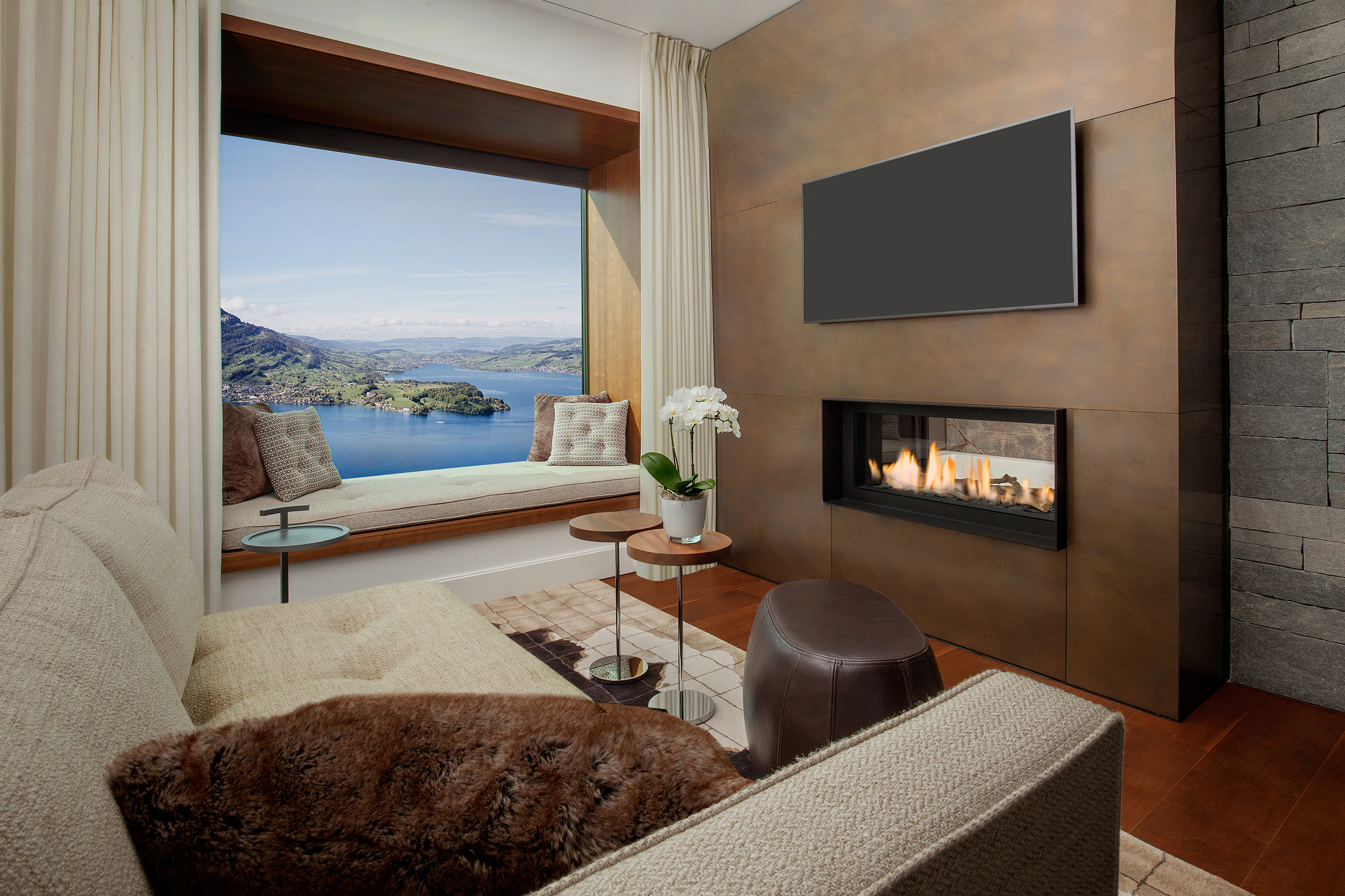A Lake View Bay Suite lounge at Bürgenstock Hotel featuring a comfy sofa in front of a fireplace with a window and windowseat overlooking the lake