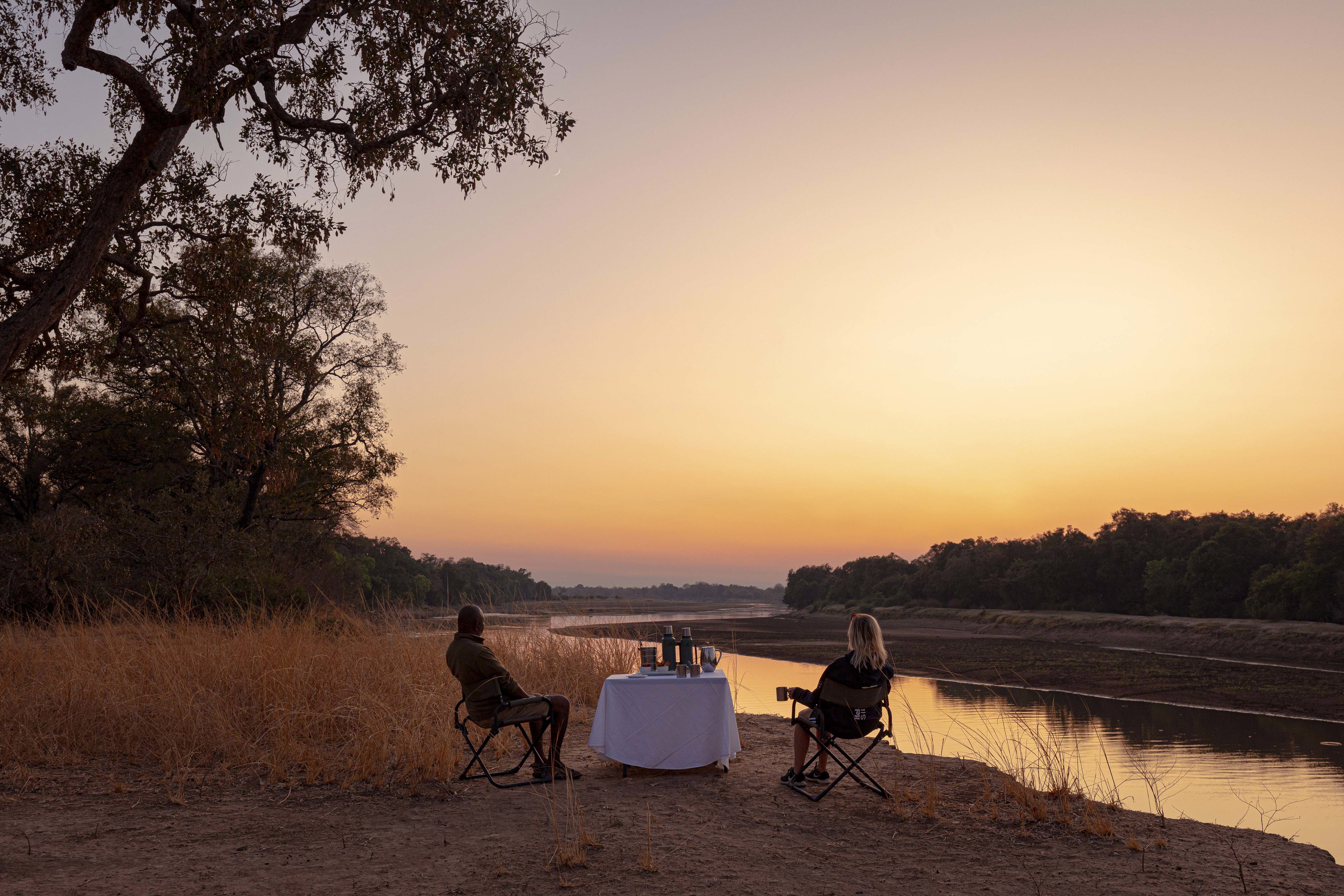 Two people enjoying a serene sunset by the Zambezi River with a table set for dining in the wilderness.