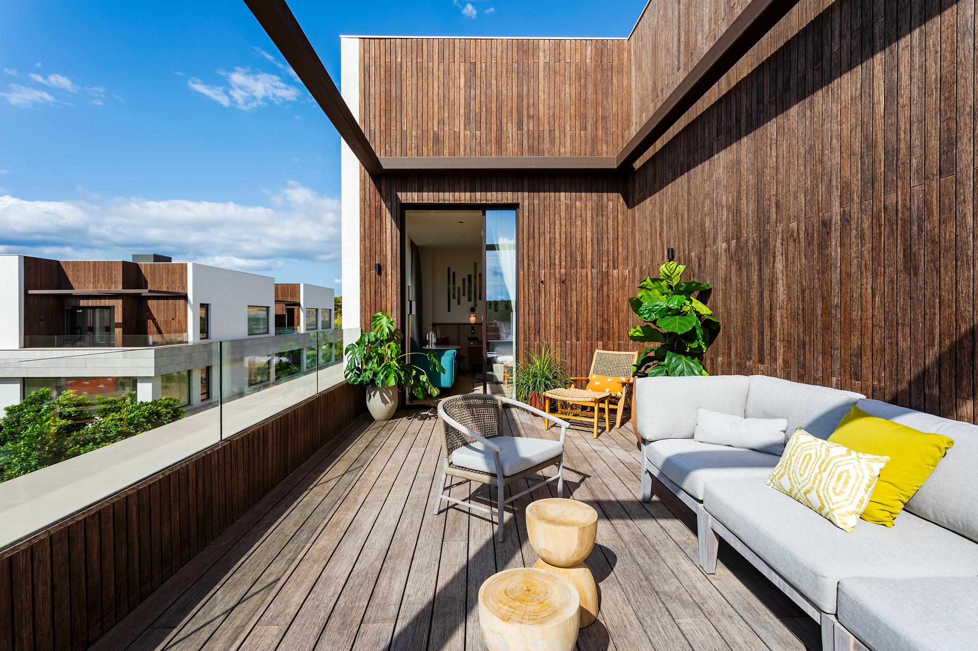 A modern terrace with wooden flooring and walls, featuring a grey sofa with yellow pillows, a wooden table, chairs, and potted plants under a clear blue sky