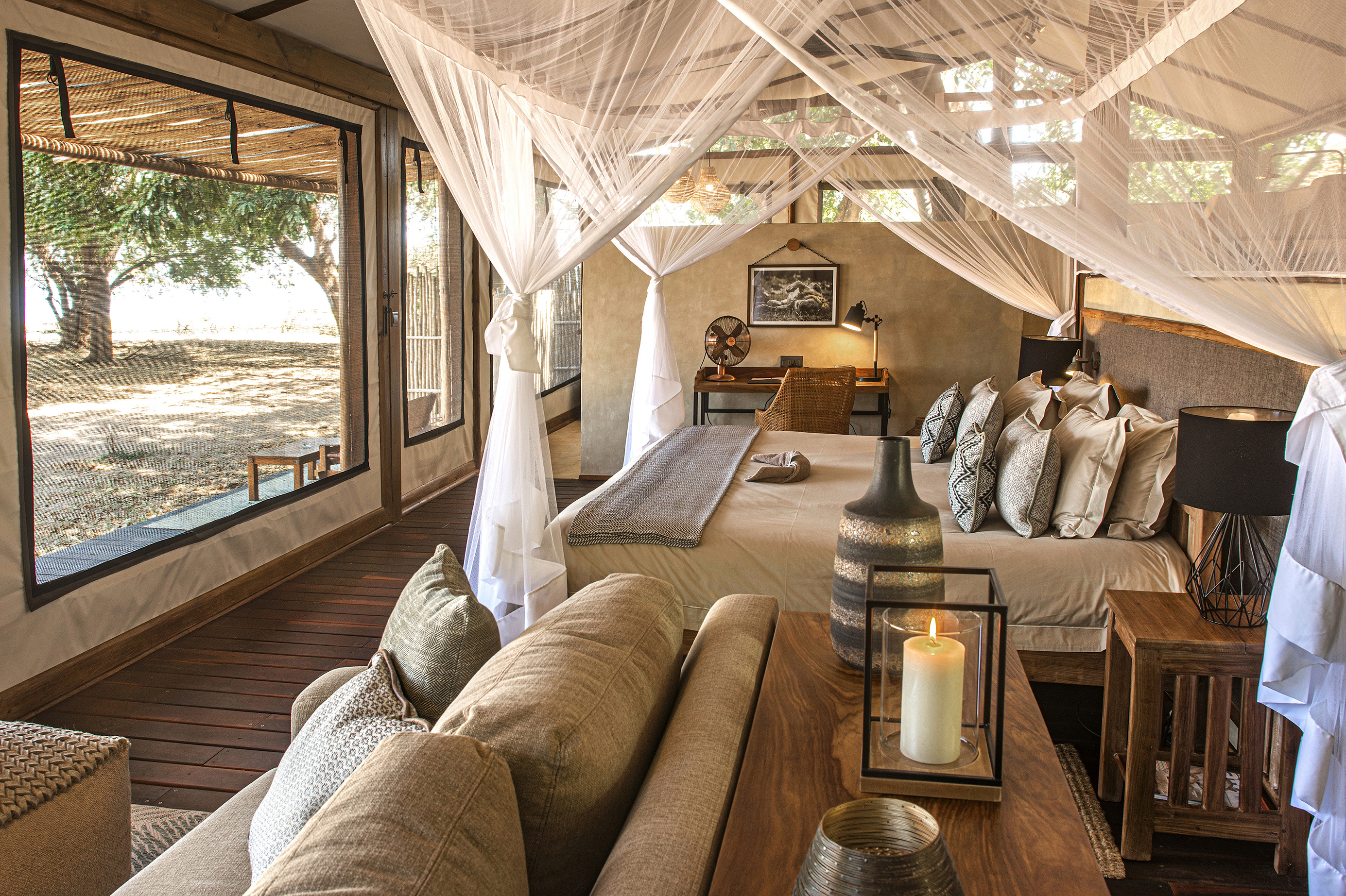The inside of a suite at Chiawa Camp decorated in neutral tones featuring a four poster bed with white nets, wooden furniture and a large window looking out
