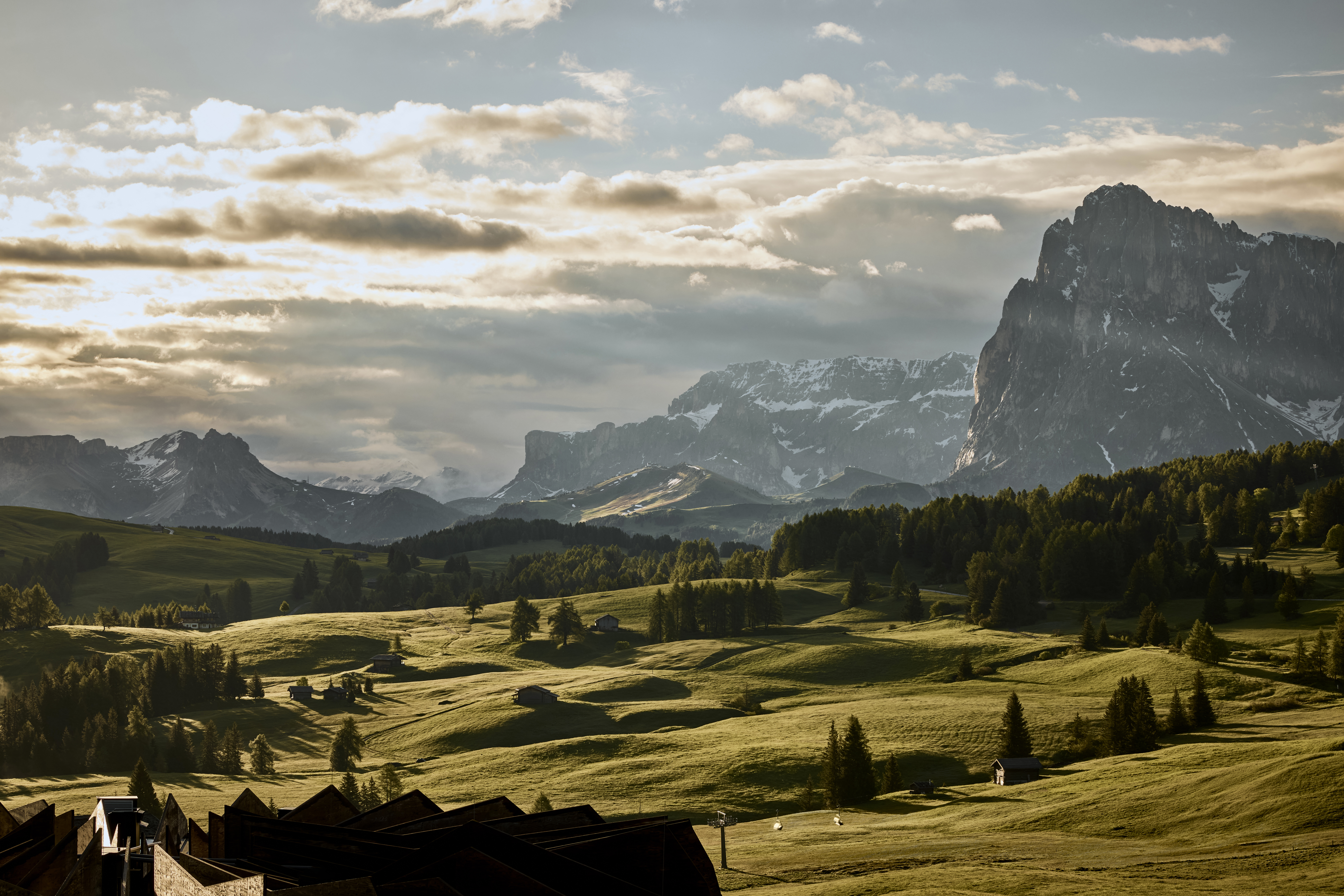 A serene landscape in the Dolomites with rolling hills, scattered trees, and a mountain range in the background under a cloudy sky.