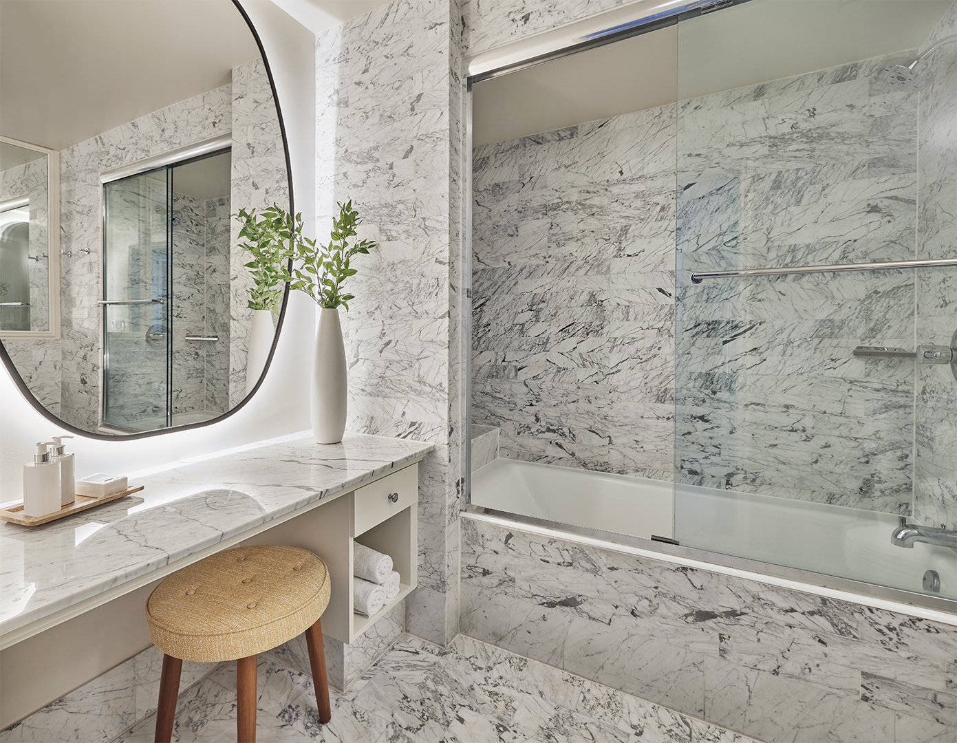 A grey marbled bathroom in a hotel suite, featuring a large tub with shower screen and a vanity area with a large lit mirror and stool.