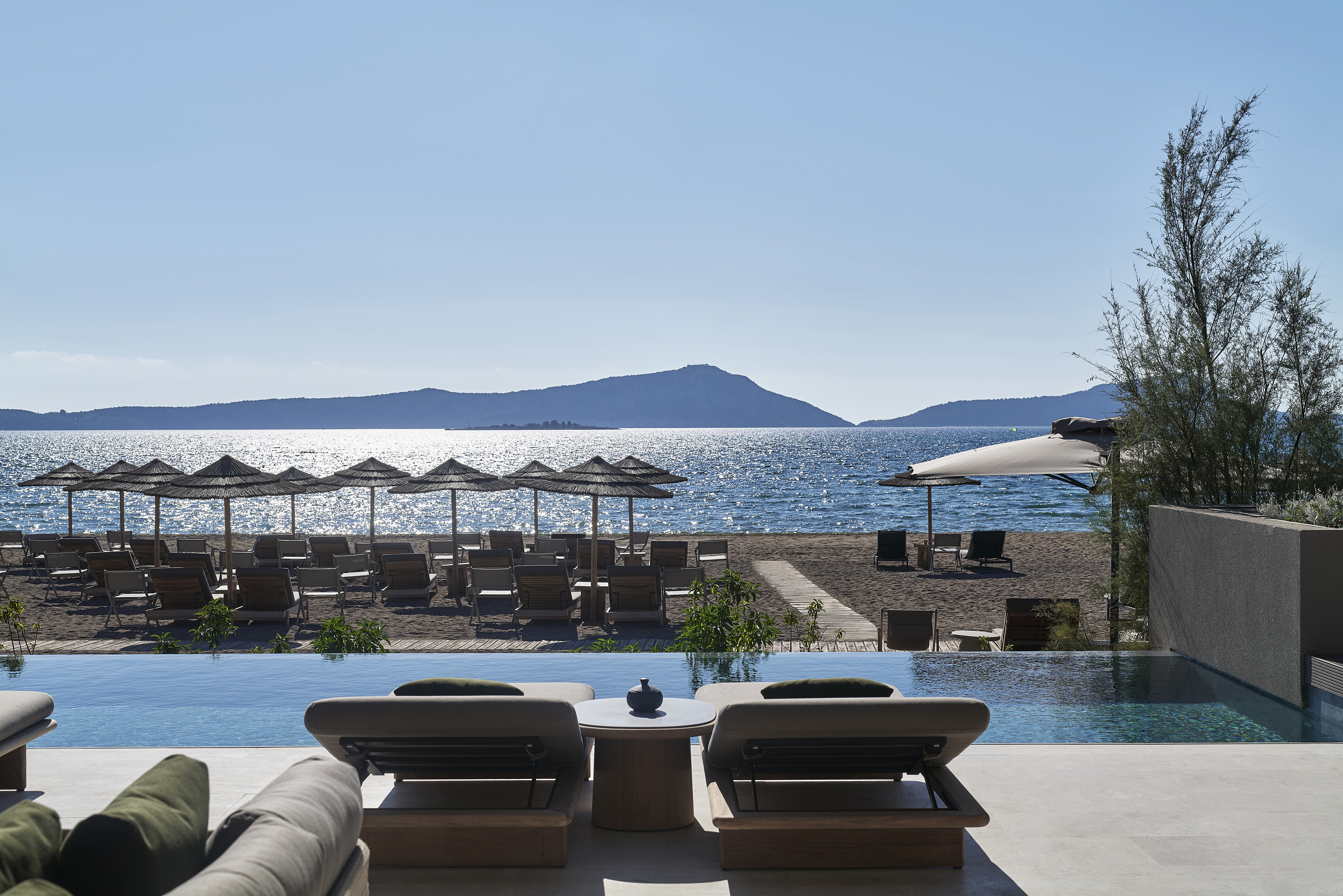 Two sun loungers facing an infinity pool looking out over a beach with more loungers and thatched umbrellas at W Costa Navarino