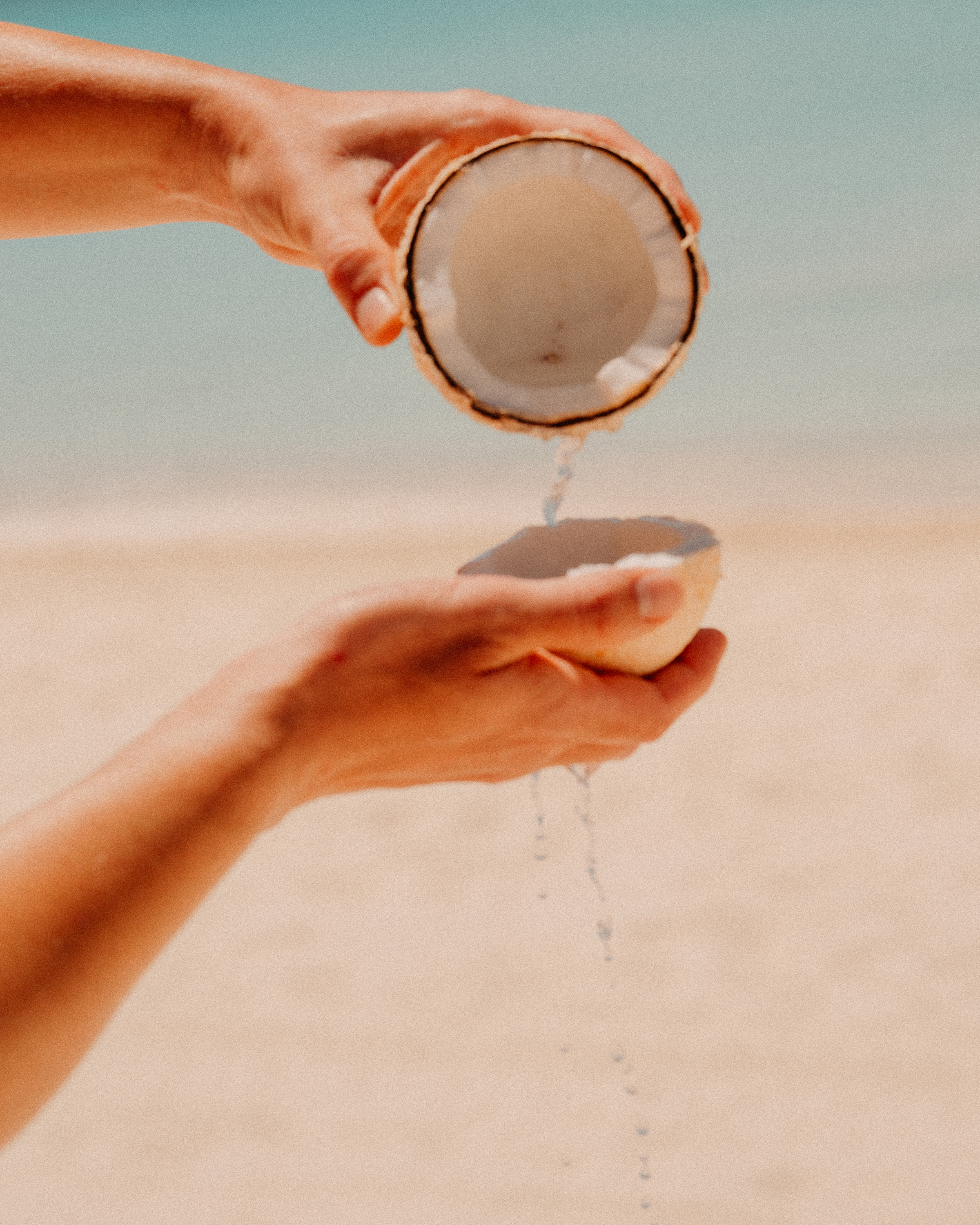 Person pouring liquid from one coconut half into another on a sandy beach with the ocean in the background