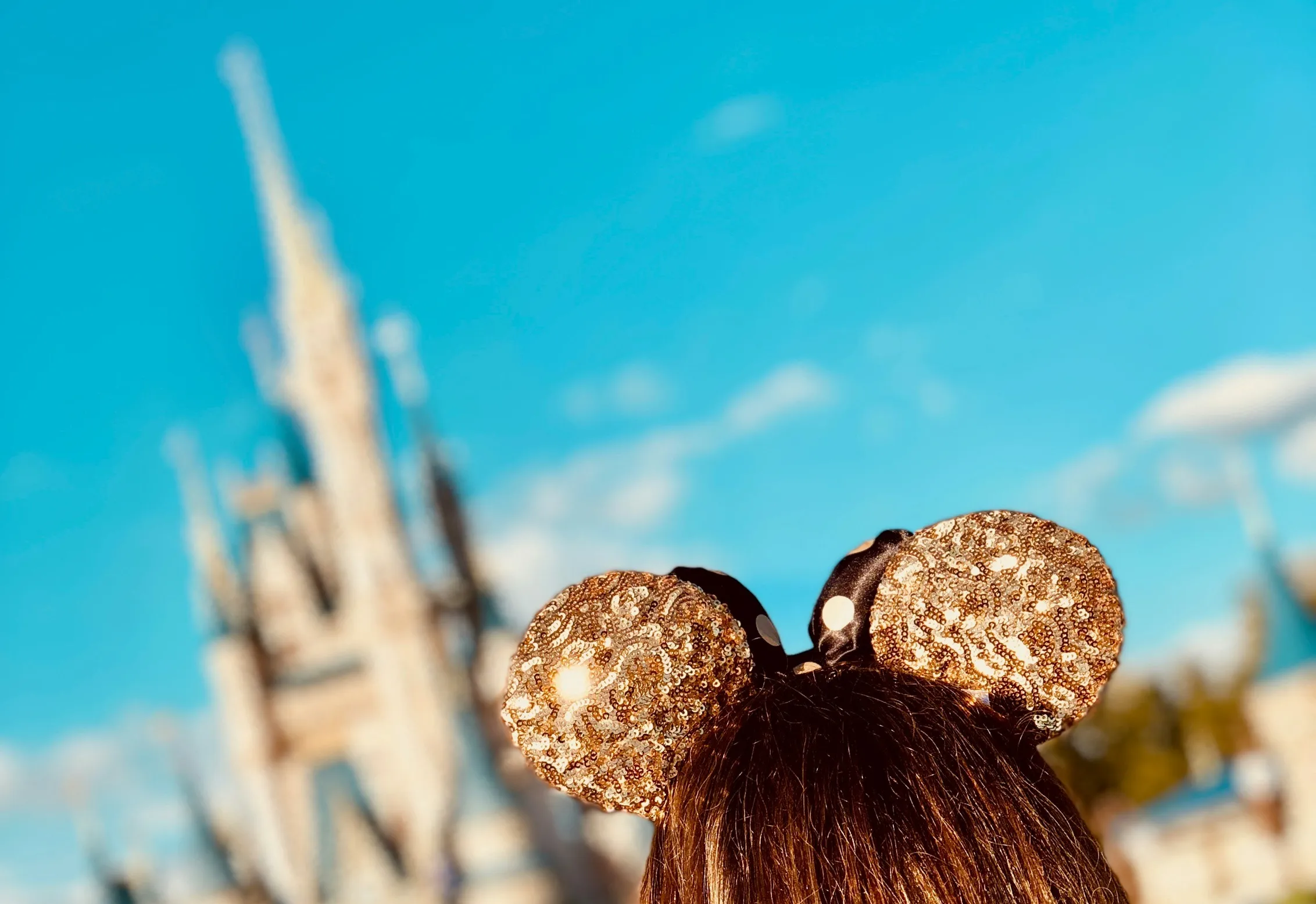 A small girl wearing glittery Mickey Mouse ears stands in front of a blurred view of Cinderella's Castle at Disney World.
