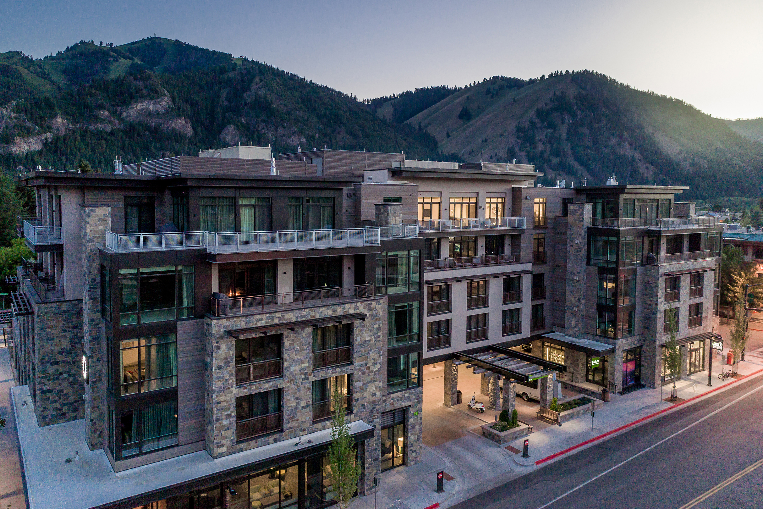 Elevated view of the Limelight Ketchum hotel at dusk, showcasing a modern multi-story building with large windows and balconies, surrounded by forested mountains