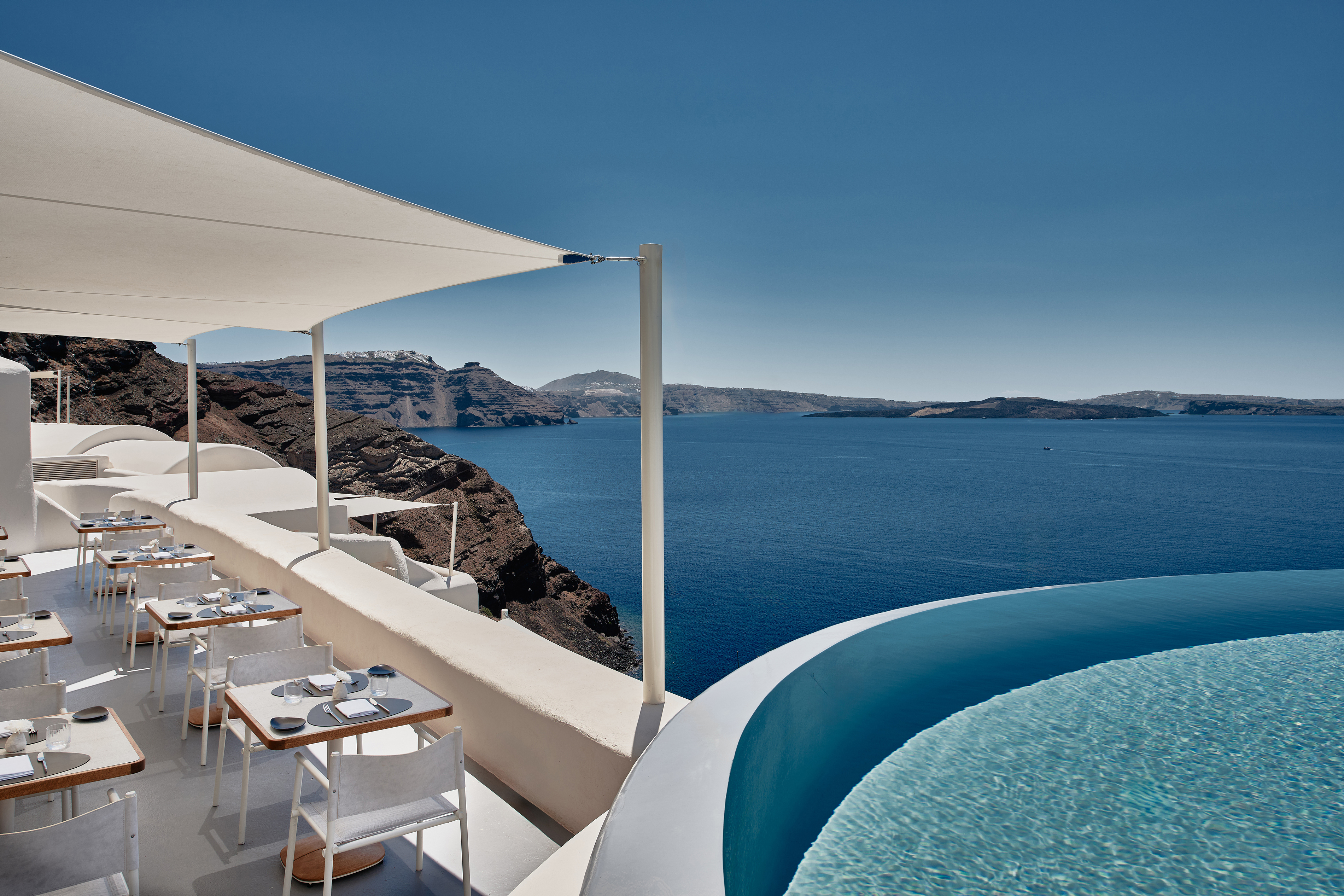 A white dining terrace with white tables and chairs covered by a canopy on a cliff edge facing the ocean, with a glimpse of a circular pool at its side