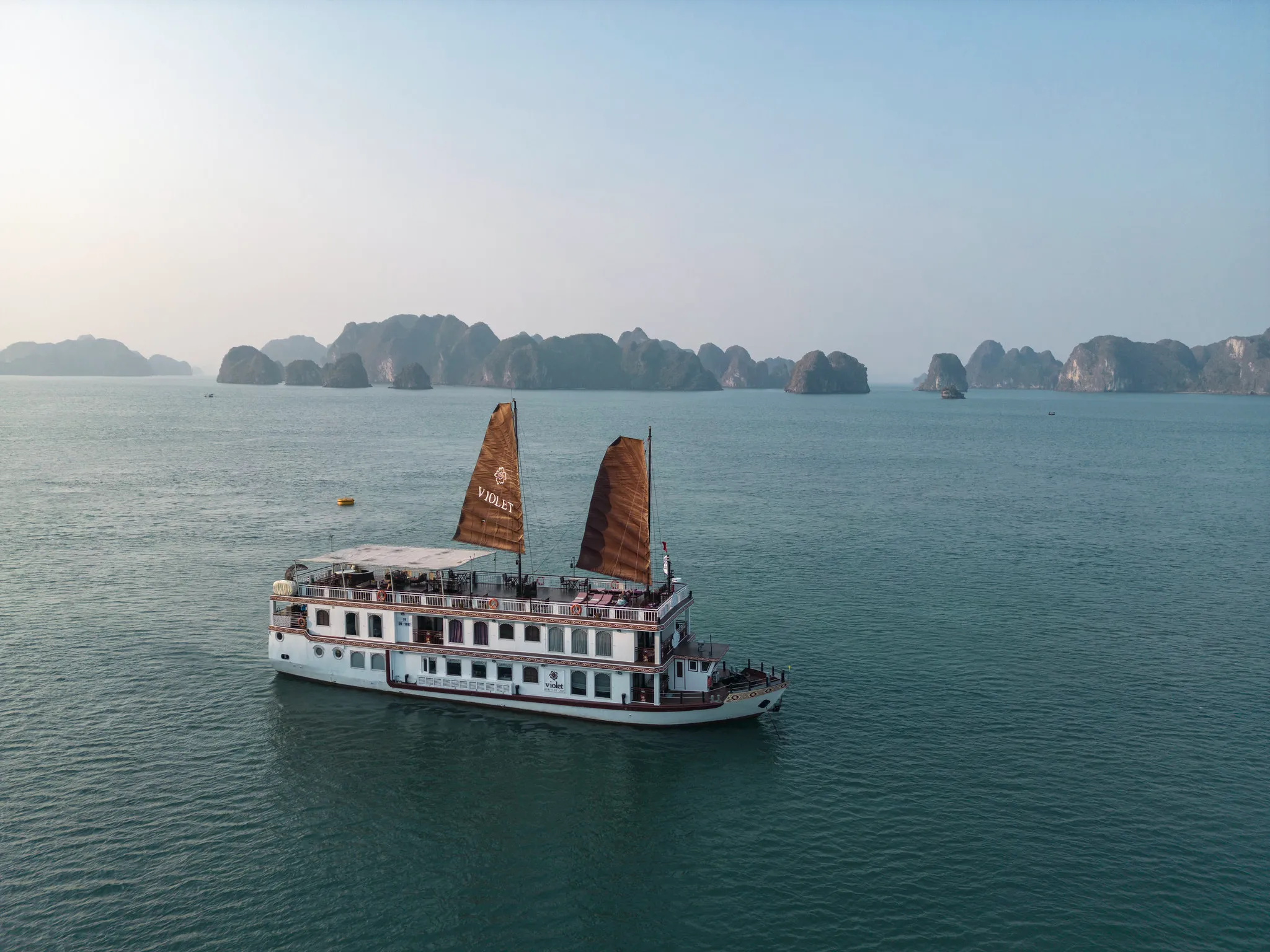 A white Heritage Line cruise ship 'Violet' with brown sails sails through calm waters with rocky islands in the background.