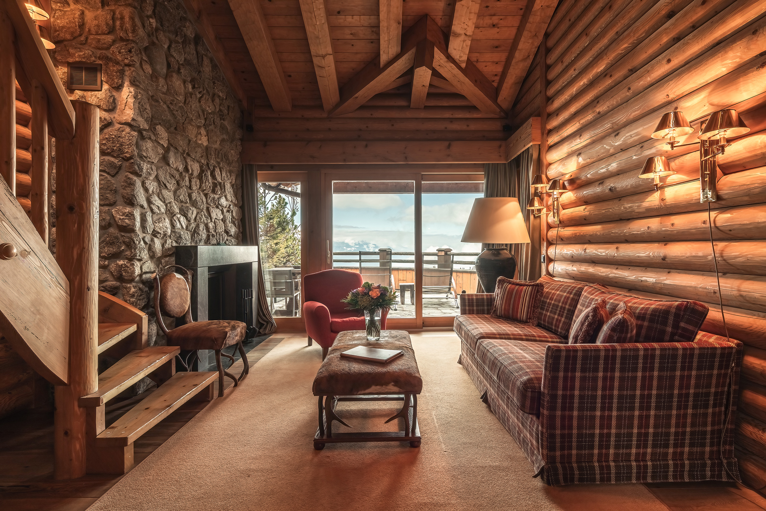 The lounge of LeCrans Prestige Suite featuring log and stone walls with comfy tarten furniture, a fireplace and sliding doors out to a balcony 