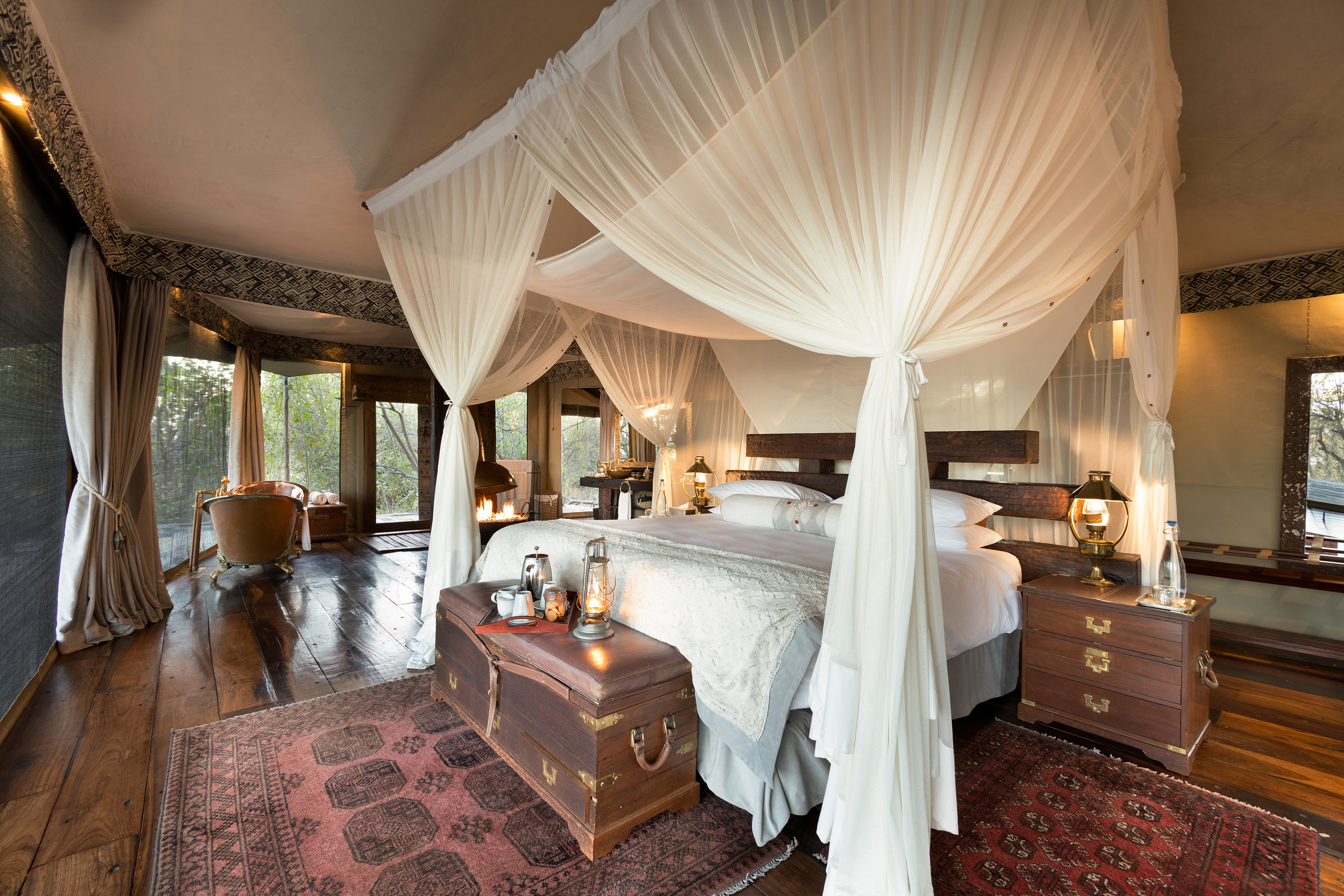 Four poster bed with white curtains in a large room decorated with trunks and a copper bath