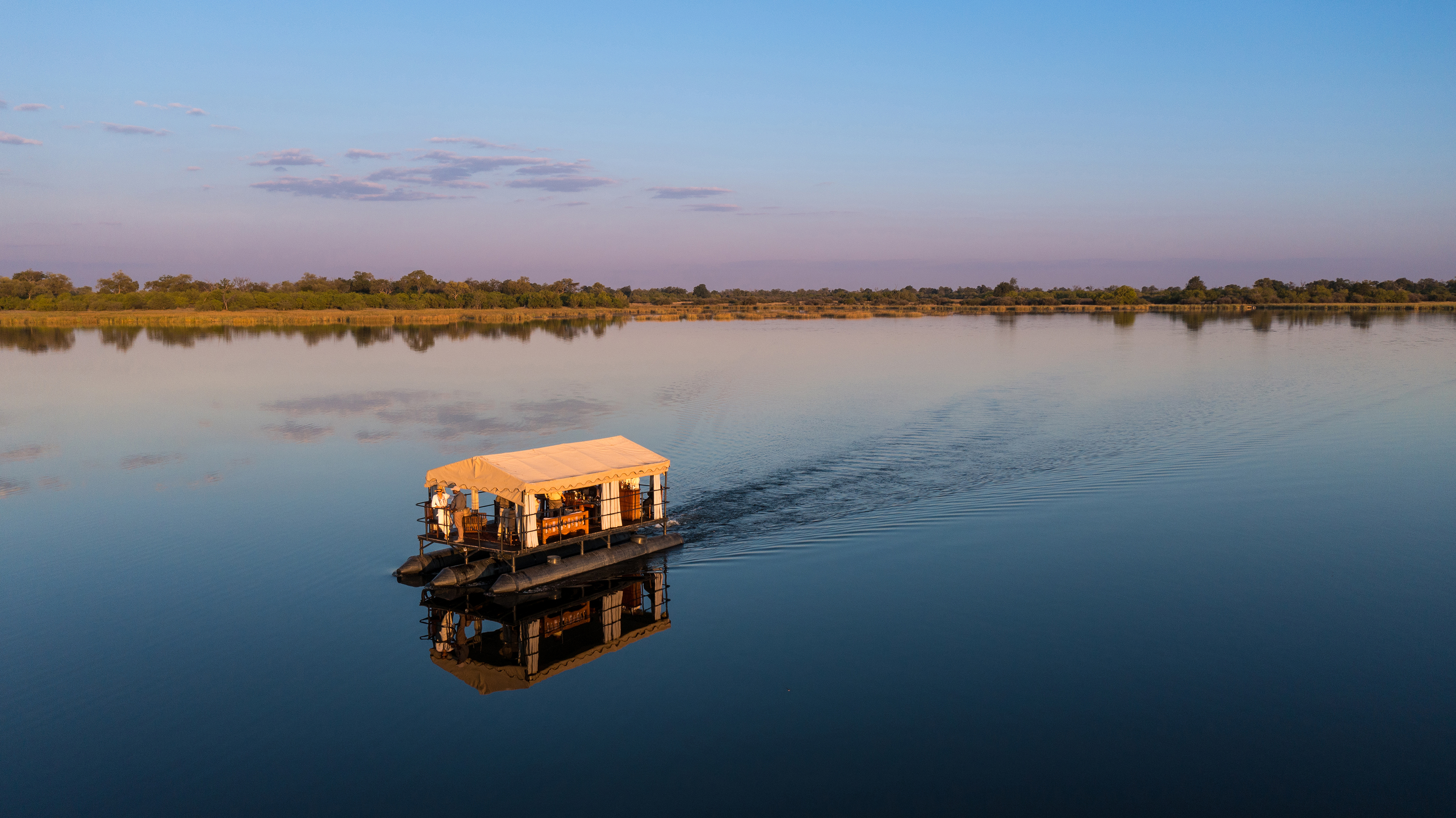 Two people standing at the front of a large motor-powered boat drifting across calm water at sunset with the African savannah on the horizon