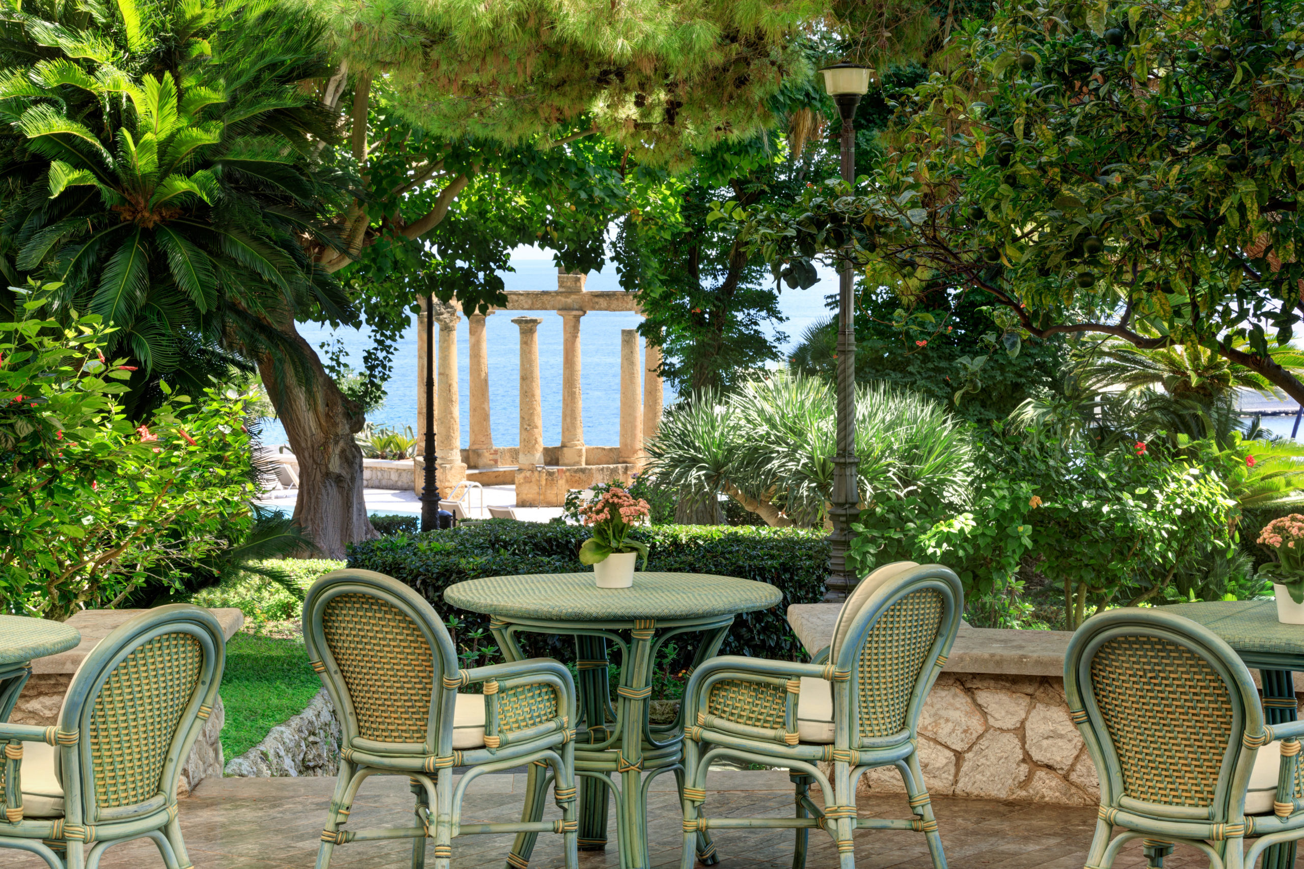 Outdoor restaurant area with green chairs and tables, surrounded by lush plants and trees, with a view of ancient stone columns in the background.