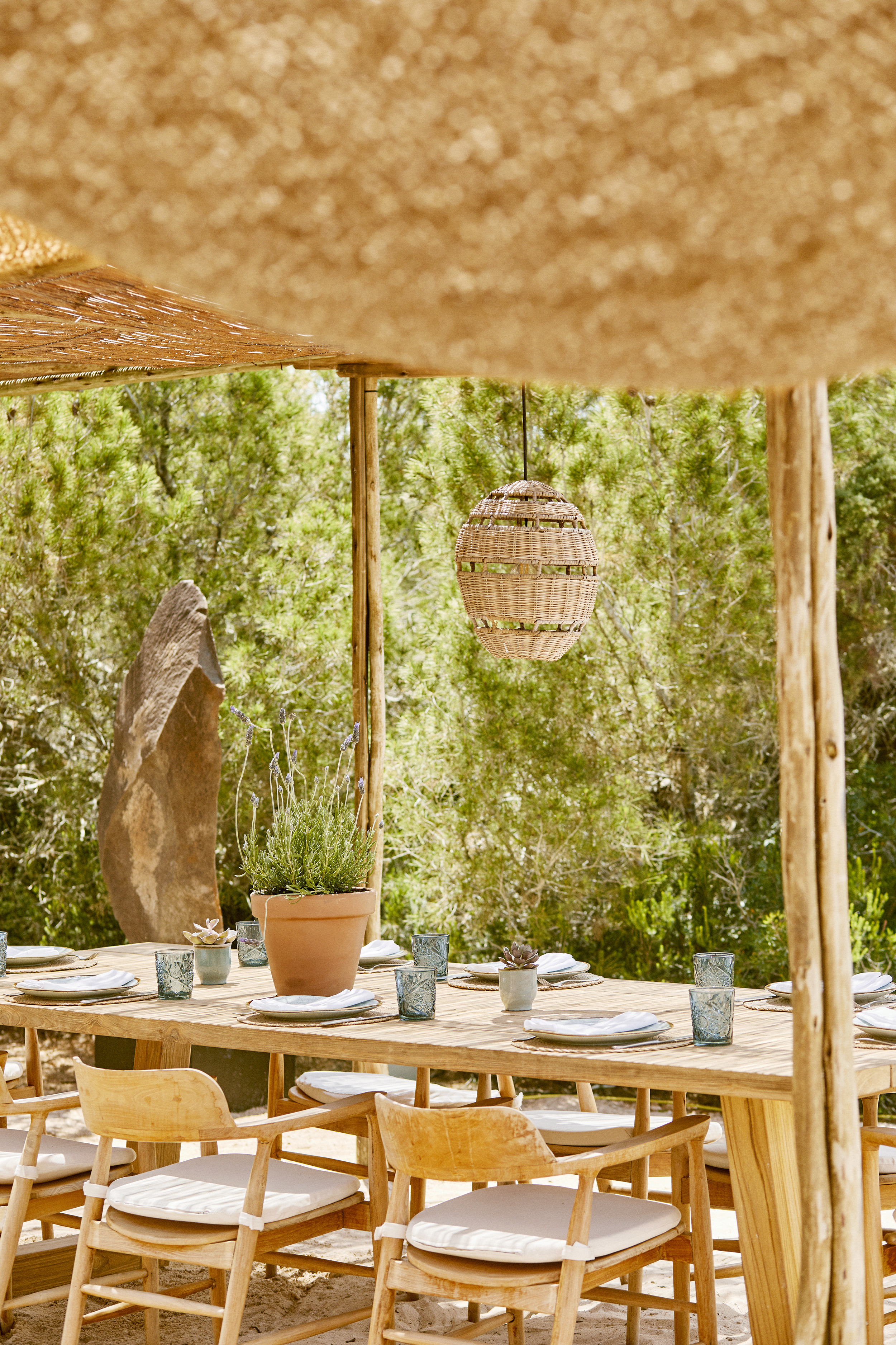 A natural wooden dining table set for dinner with blue glassware and a terracotta plant pot beneath a hessian canopy with trees behind