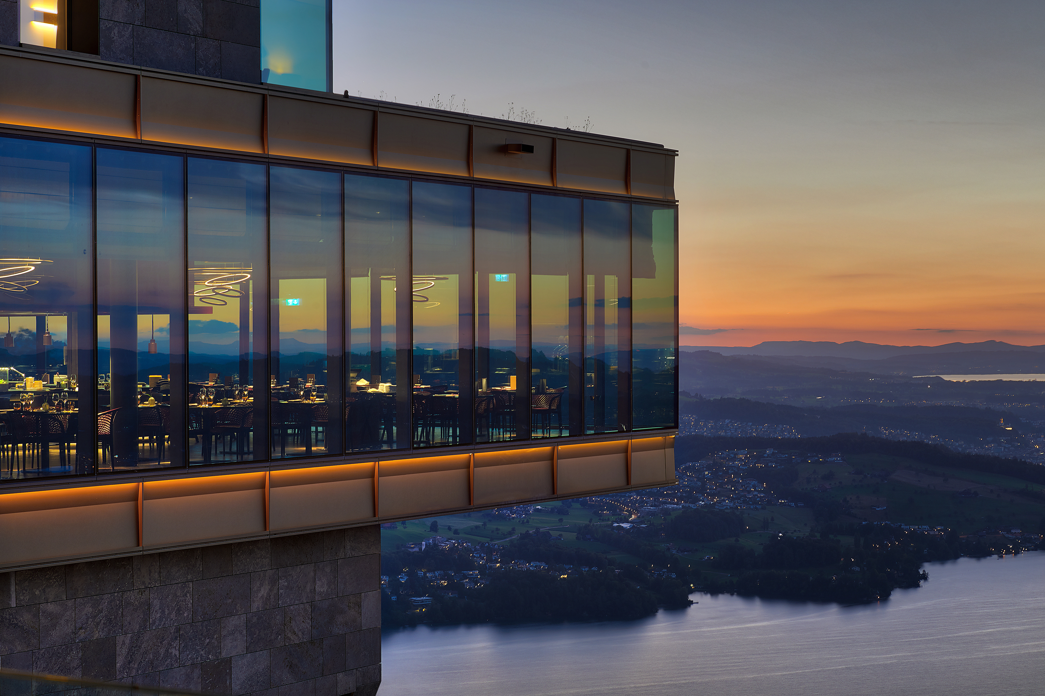 A modern restaurant with floor to ceiling windows that partially hangs high above a lake at sunset