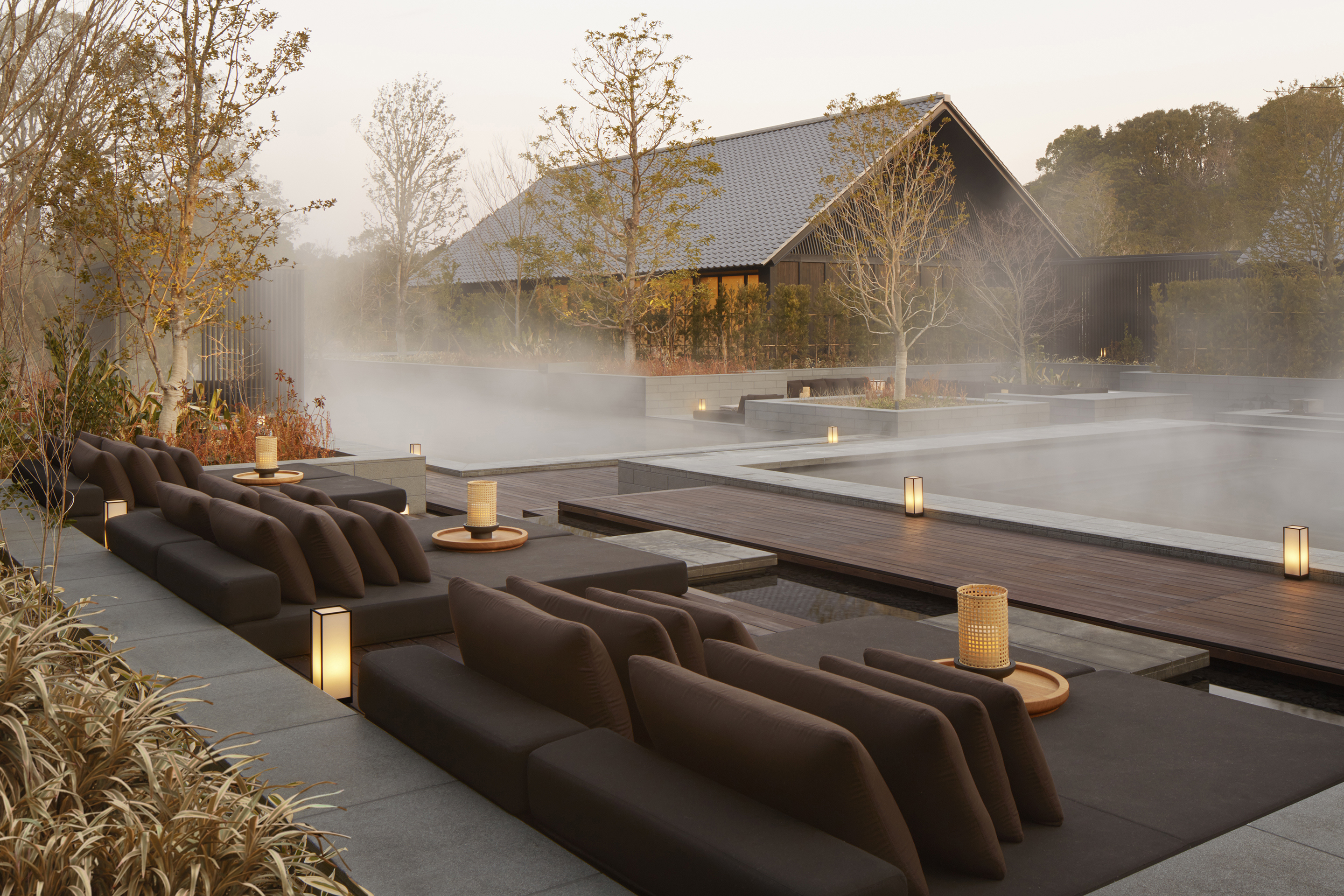 The steaming outdoor onsen at Amanemu surrounded by trees, dark sun loungers and lit candles with a view of the hotel through the steam