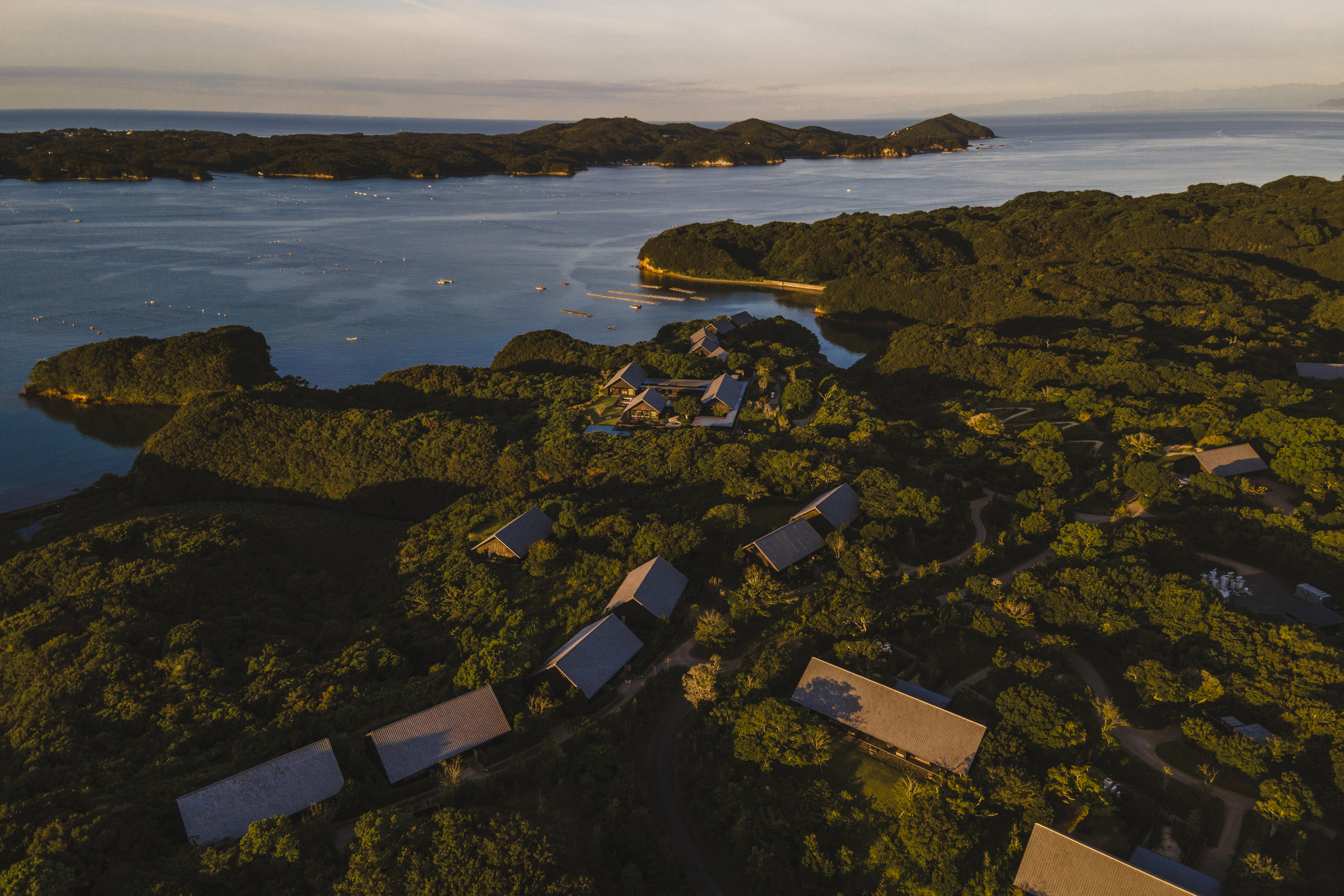 An aerial view of Amanemu featuring the roofs of the hotel buildings among the jungle with a view of the ocean and other land beyond