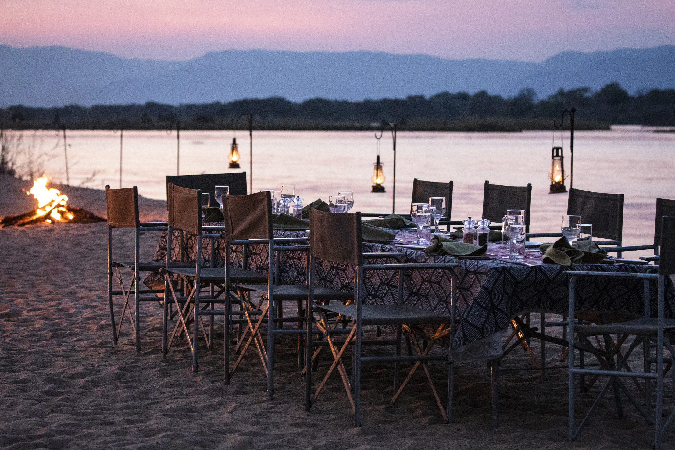 An outdoor dinner setting featuring a table and chairs set in the sand beside the Zambezi River beside a bonfire at dusk