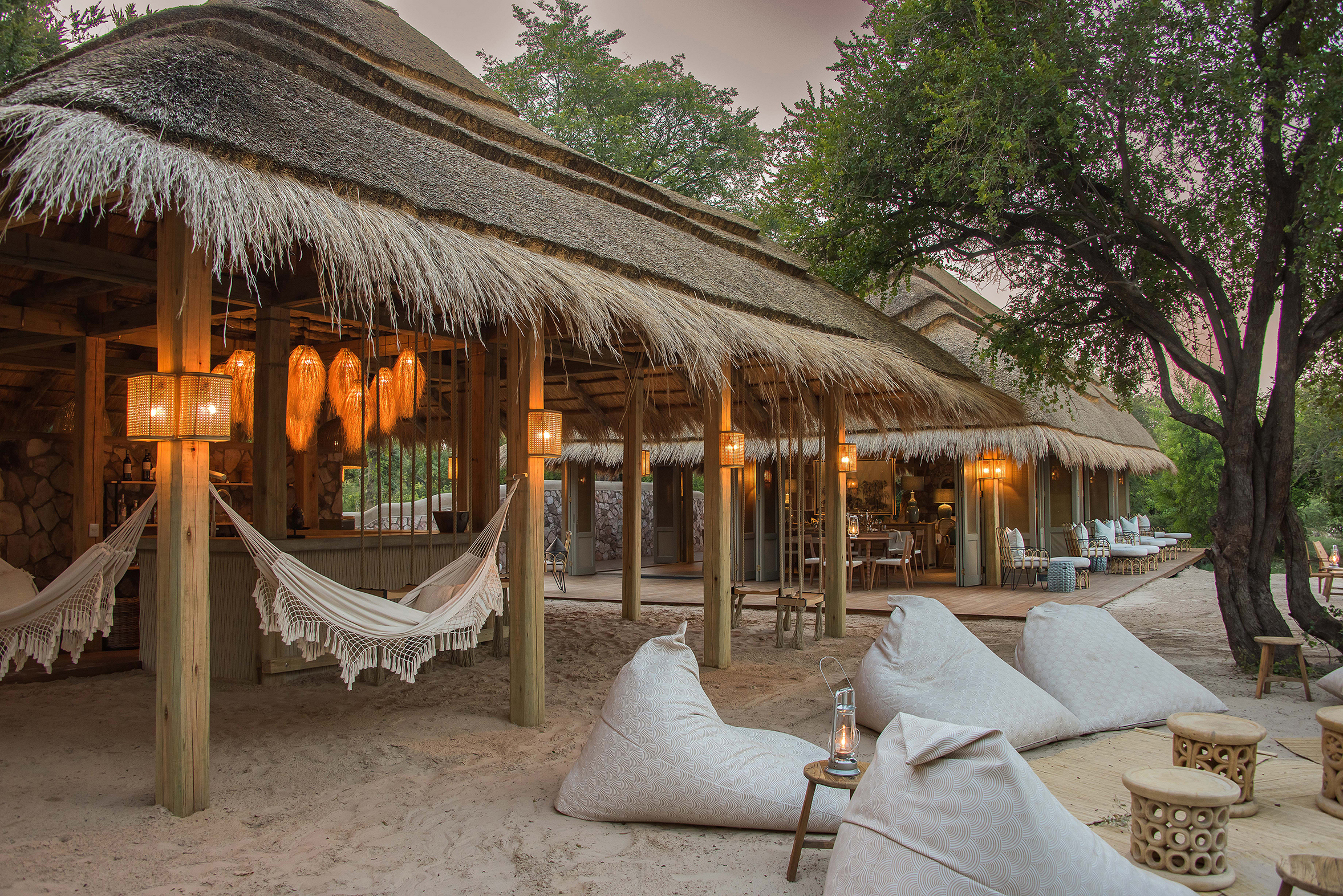 An outdoor area with comfy bean bag seats and a bar beneath a thatched roof featuring hammocks and swinging chairs