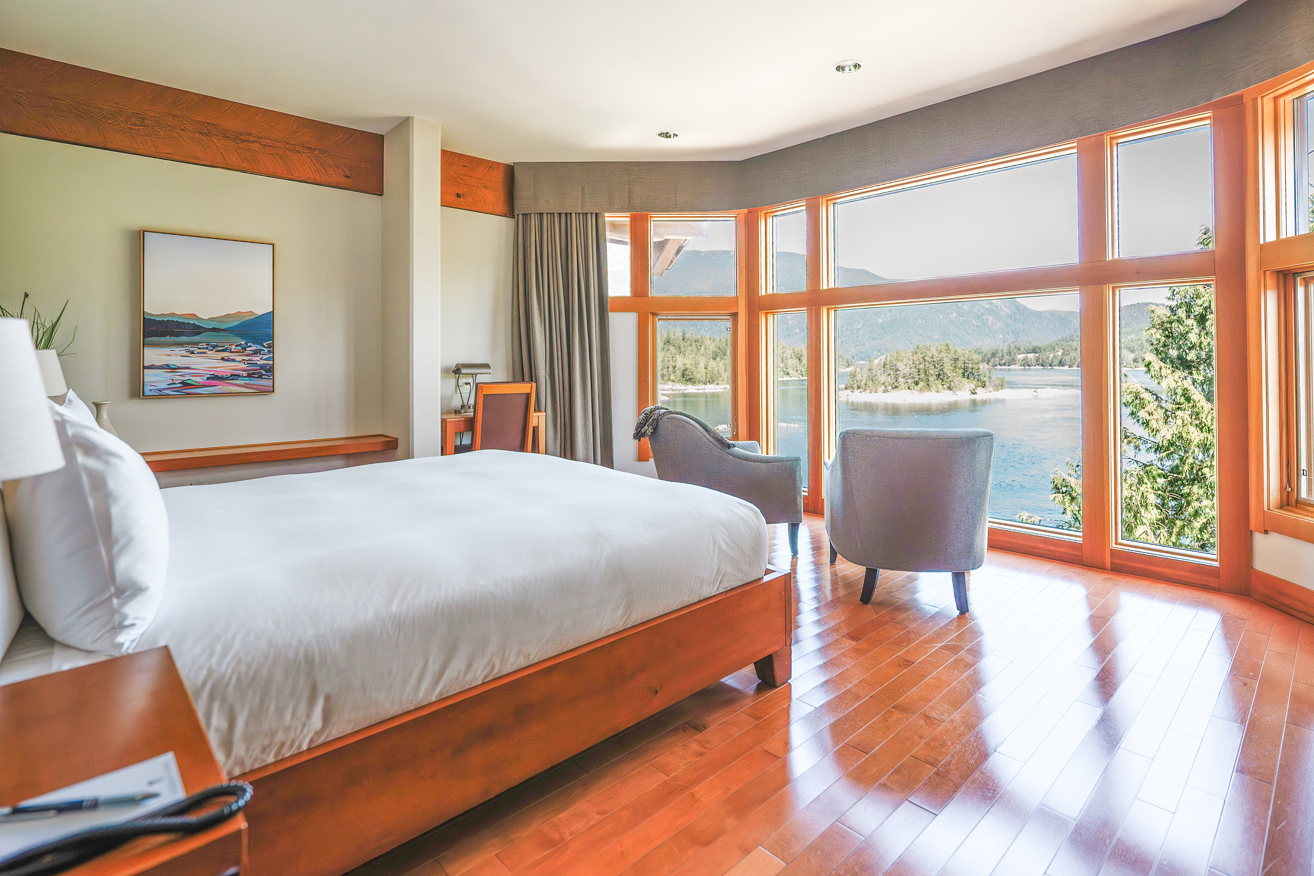 A cosy bedroom with a bed and two chairs facing wrap-around windows with a view of the landscape